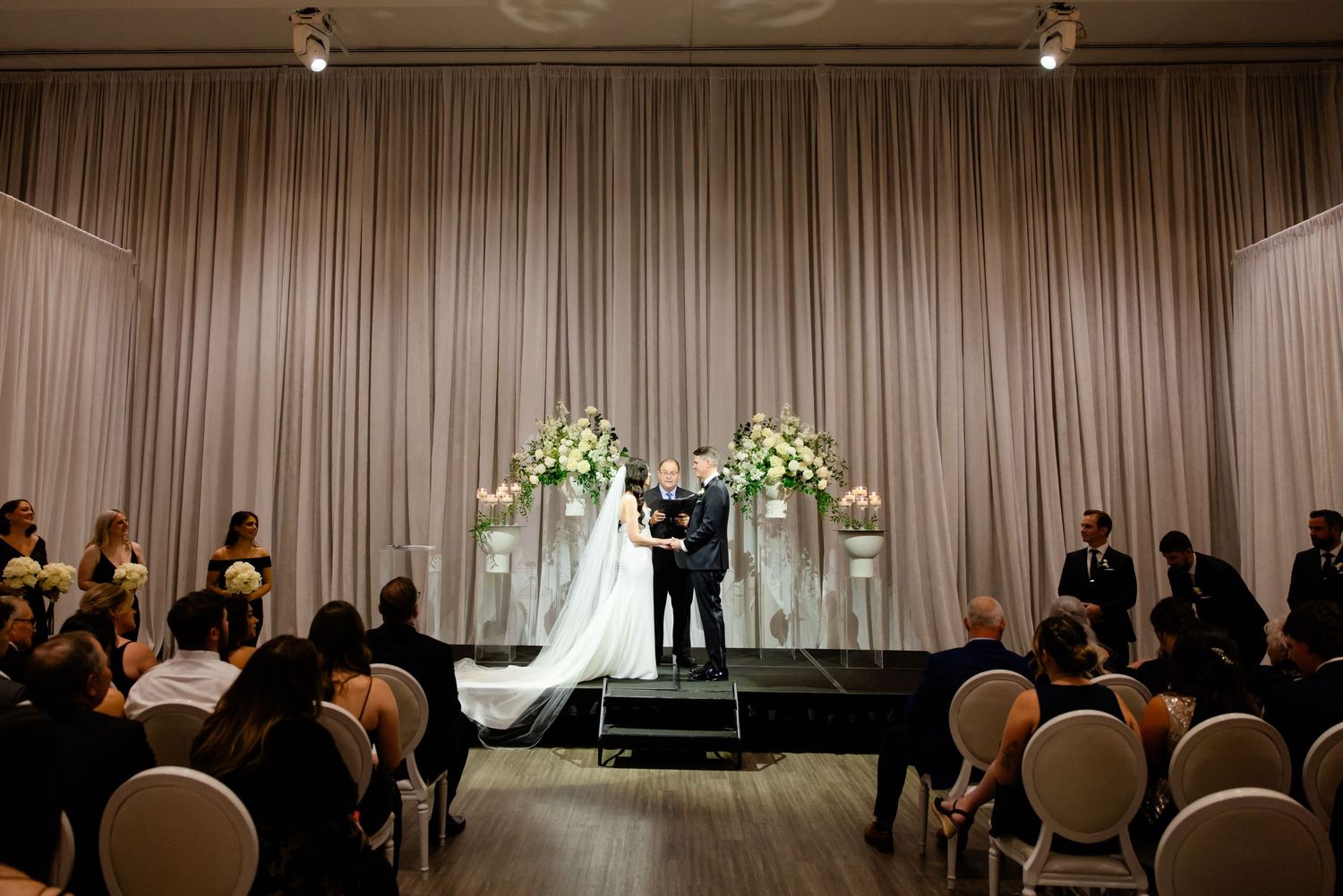 Wide view of wedding ceremony with guests seated in white chairs facing decorated stage.