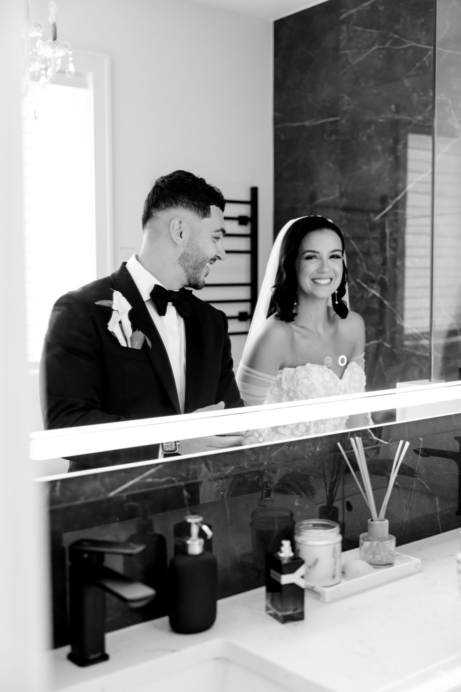 Wedding couple smiling at each other in a bathroom mirror reflection with modern fixtures.