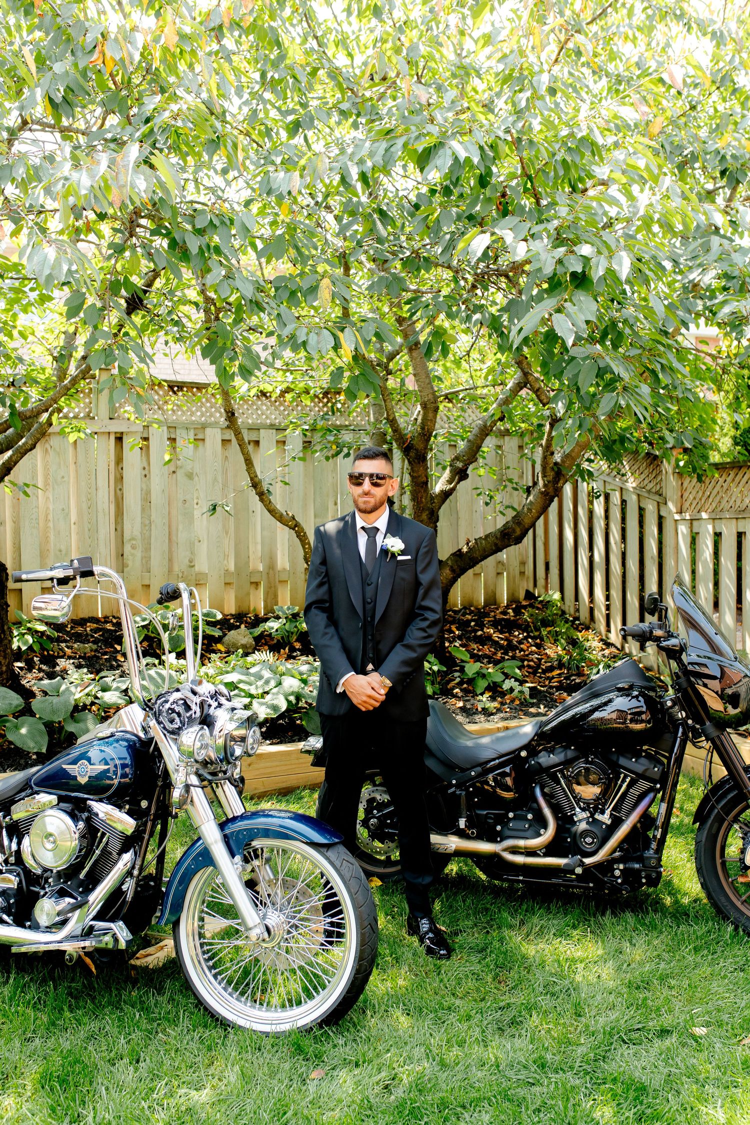 Sharp dressed figure posing between two custom motorcycles in a garden setting.
