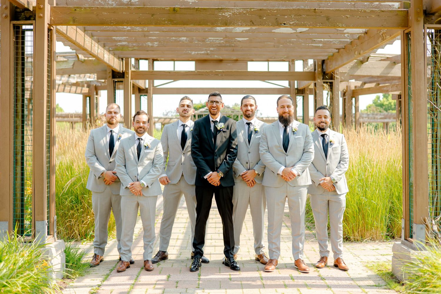 Groomsmen in light gray suits pose with groom under wooden pergola.