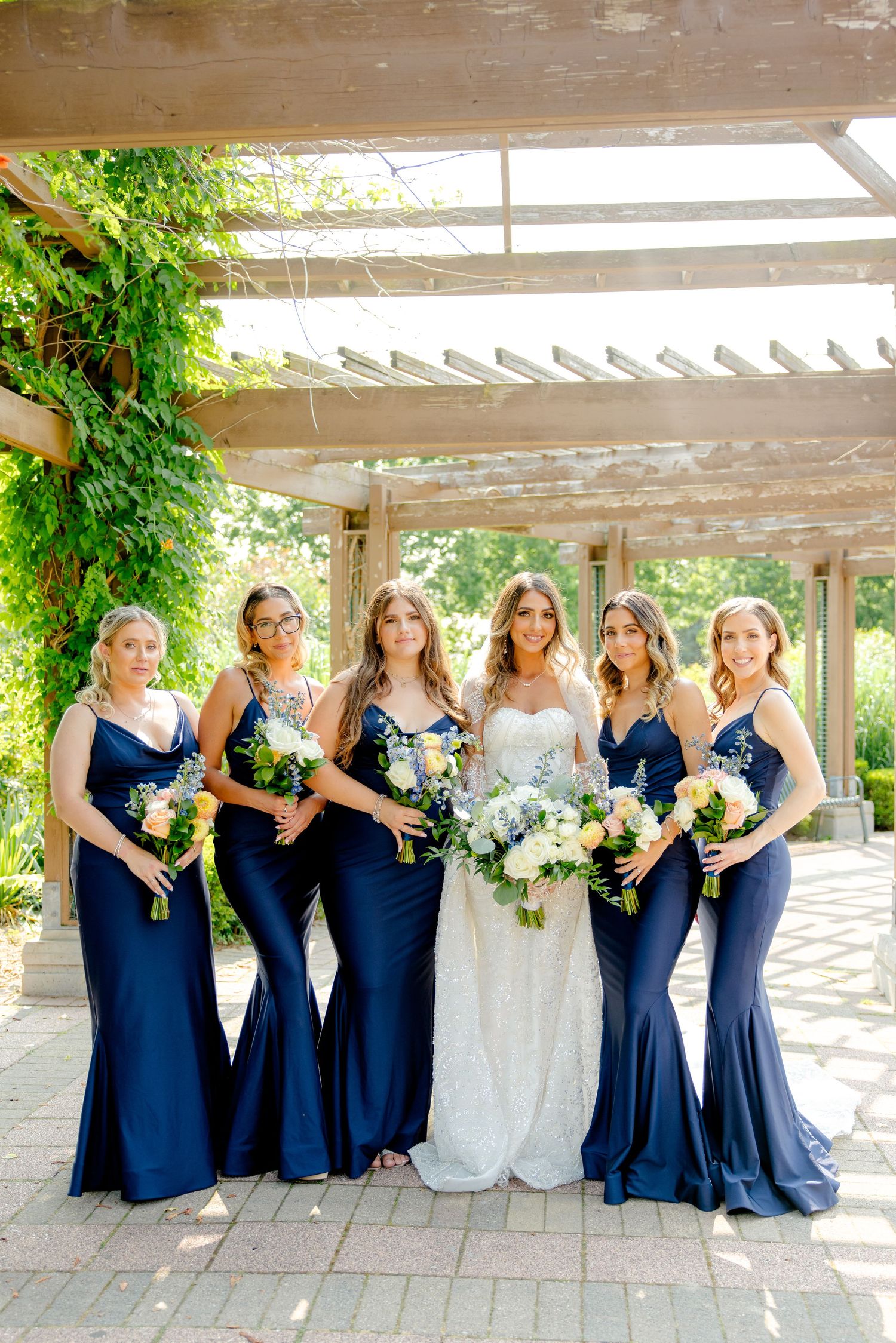 Wedding party poses under a wooden pergola with bridesmaids in navy blue dresses holding floral bouquets.