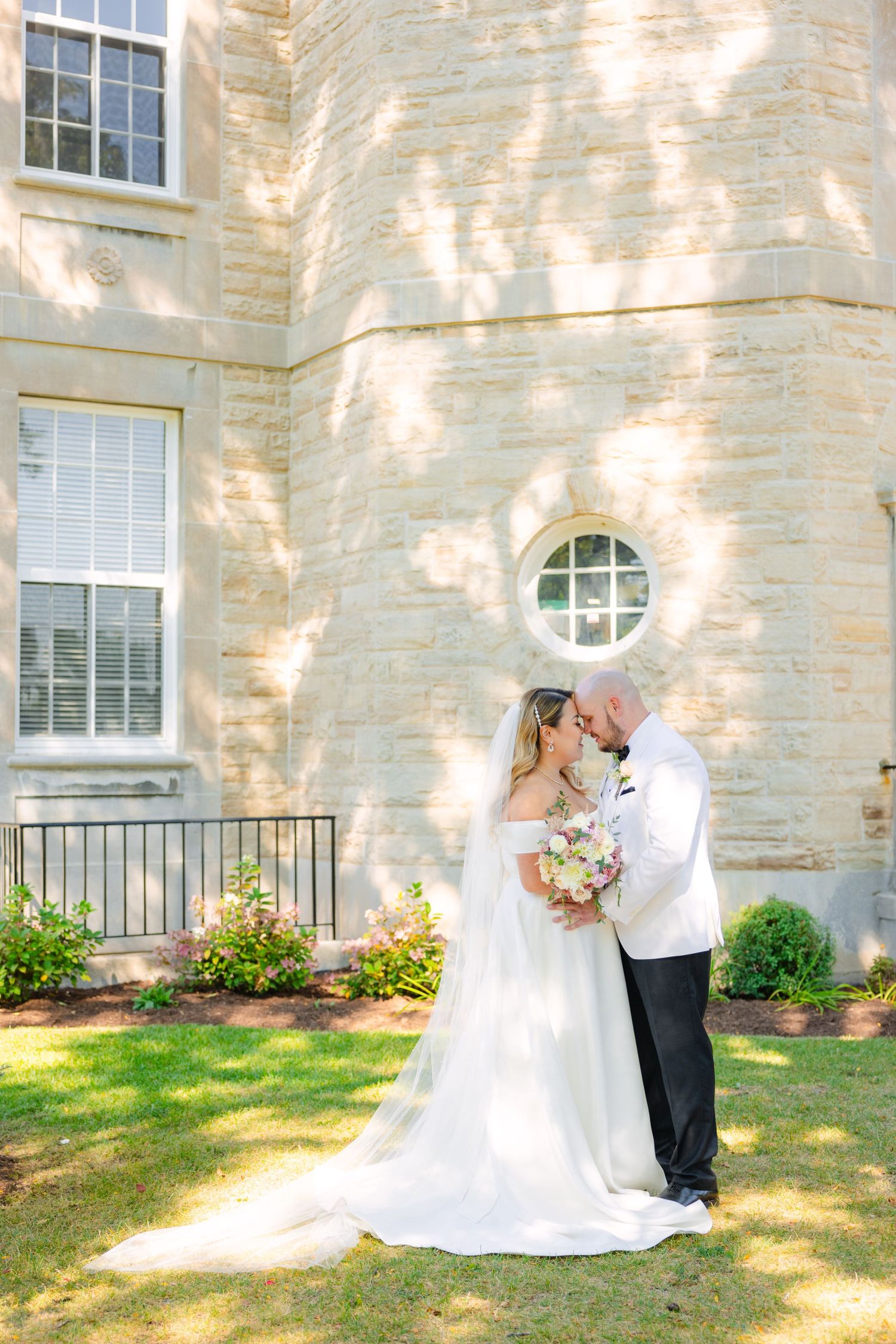 Newlyweds share tender moment in front of historic building with dappled sunlight.