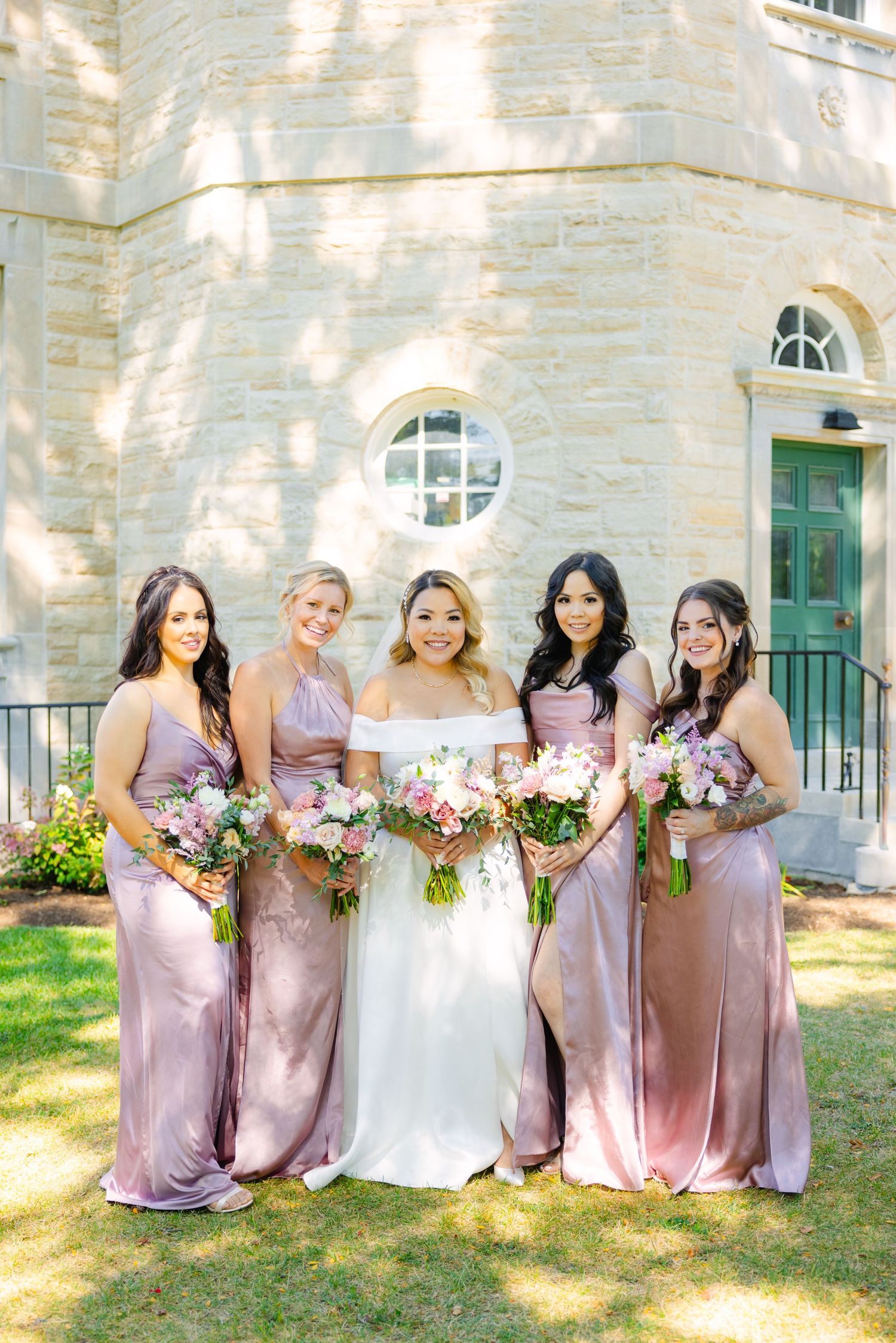 Bridesmaids in flowing pink dresses pose with bride in white gown against stone church backdrop.