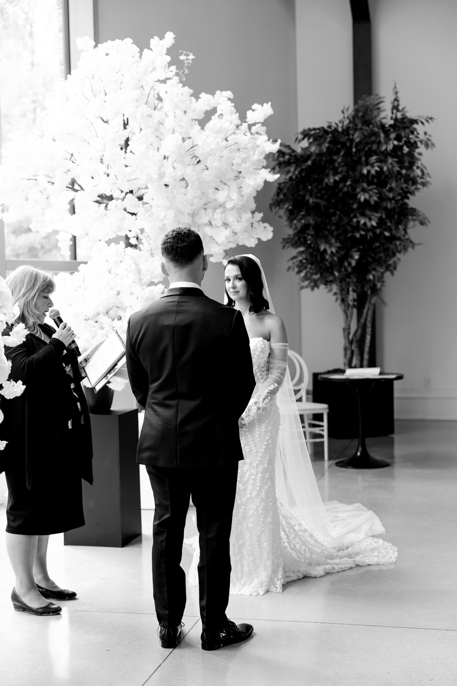 A wedding ceremony in a modern venue with dramatic white floral arrangements and natural light.