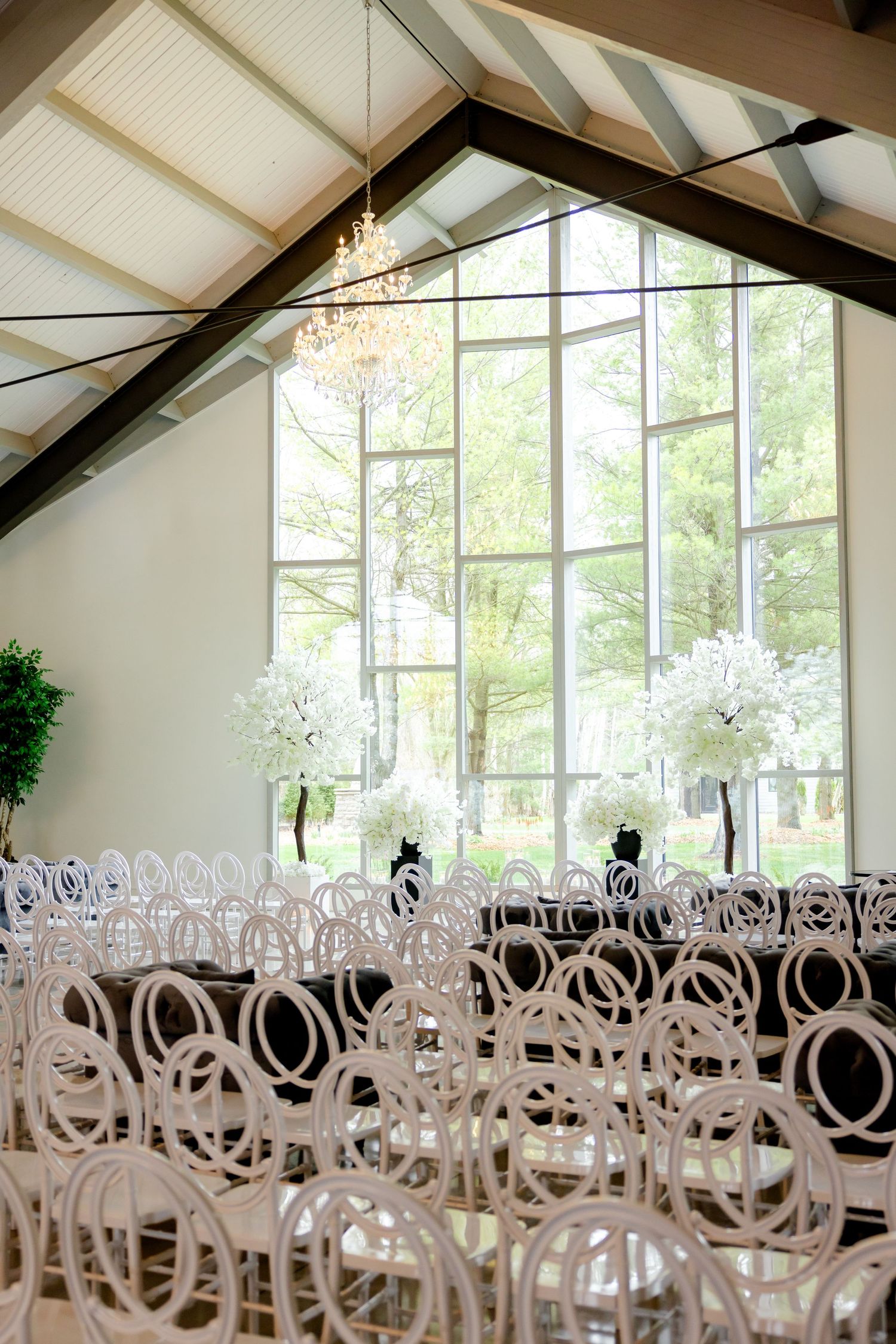 Modern wedding venue with white decor, crystal chandelier, and geometric chairs in front of large windows.