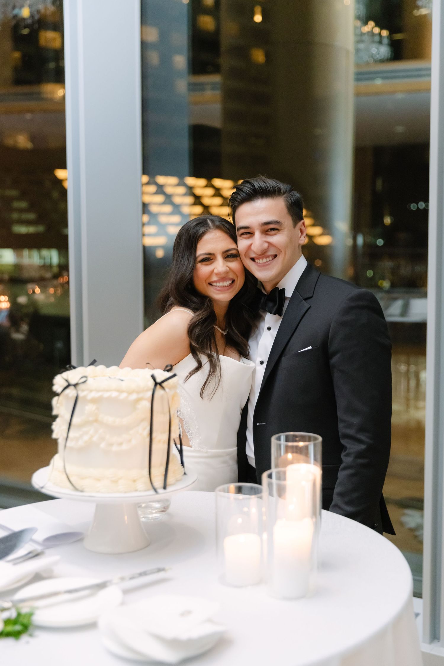 A couple in wedding attire poses next to their elegant white wedding cake on a candlelit table.