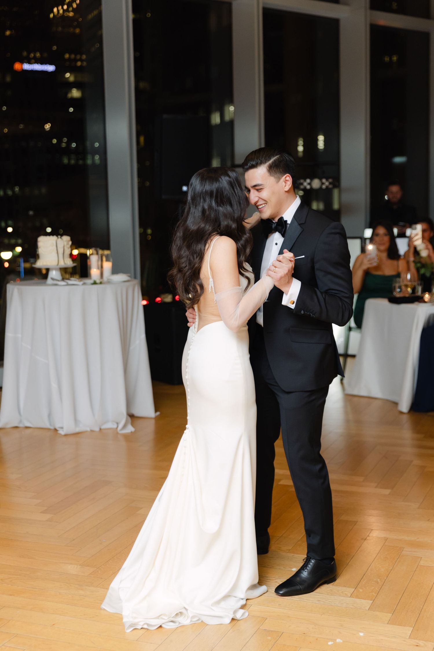 Couple shares romantic dance moment in elegant reception venue with city lights backdrop.
