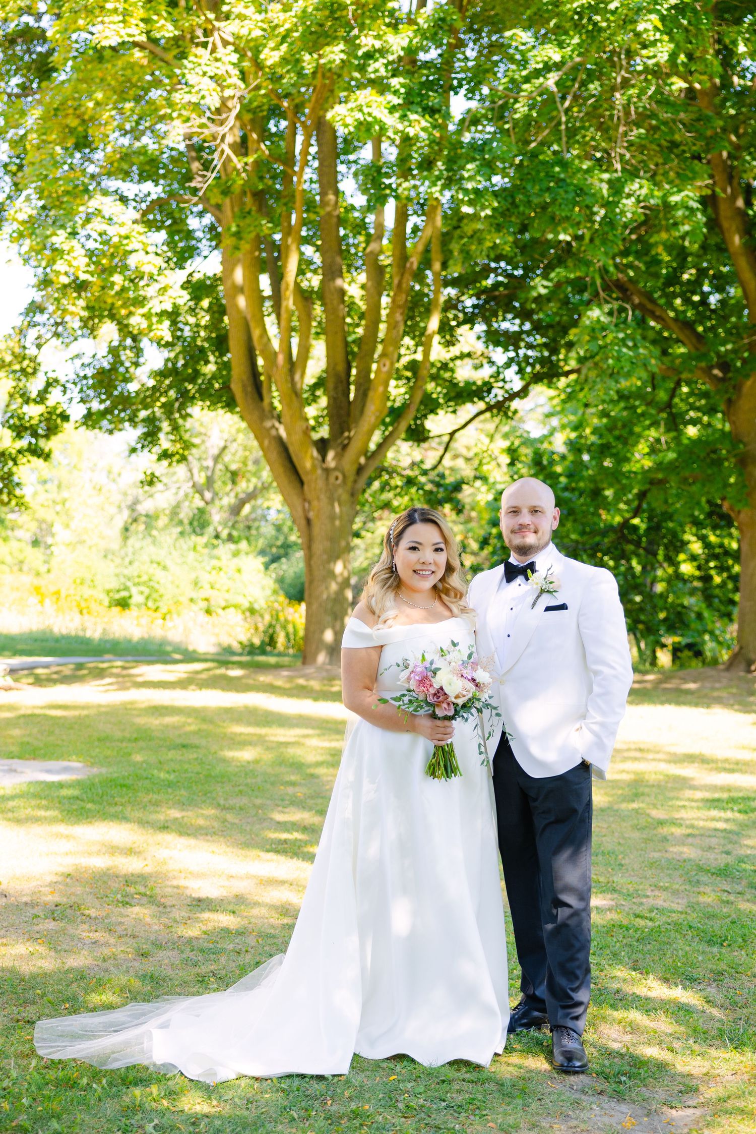 Newlyweds stand together beneath towering trees at outdoor wedding venue on sunny day.