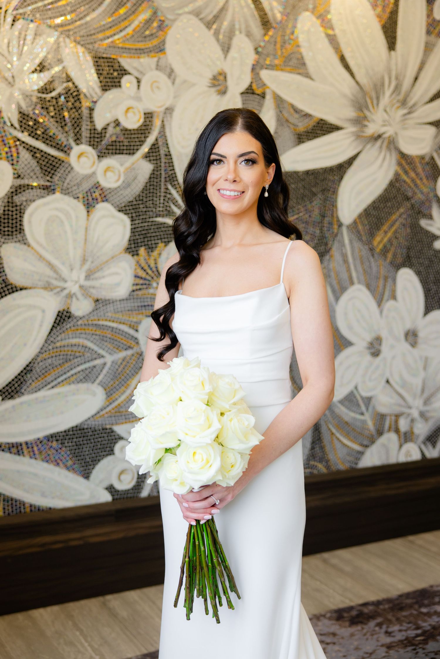 Elegant bride holding white rose bouquet against metallic floral wallpaper.