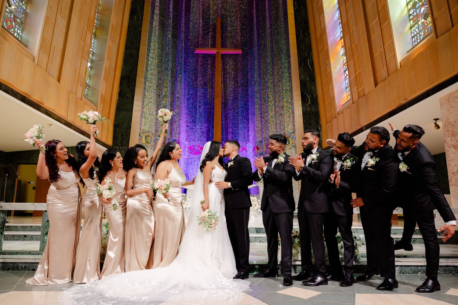 Joyful wedding party celebrates with raised arms at church altar under purple lighting.