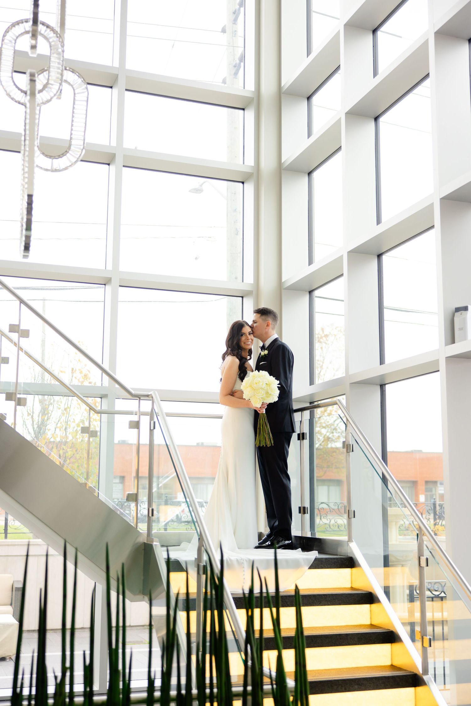 Wedding photograph featuring couple on illuminated yellow stairs with glass walls and decorative plants.