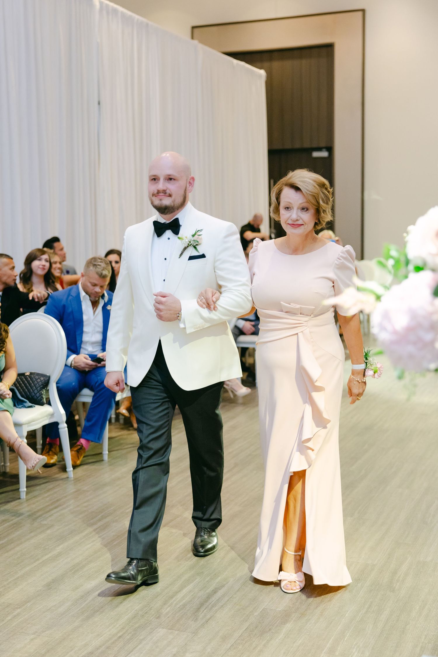 A formal processional walk featuring a couple arm-in-arm in a modern wedding venue with white decor.