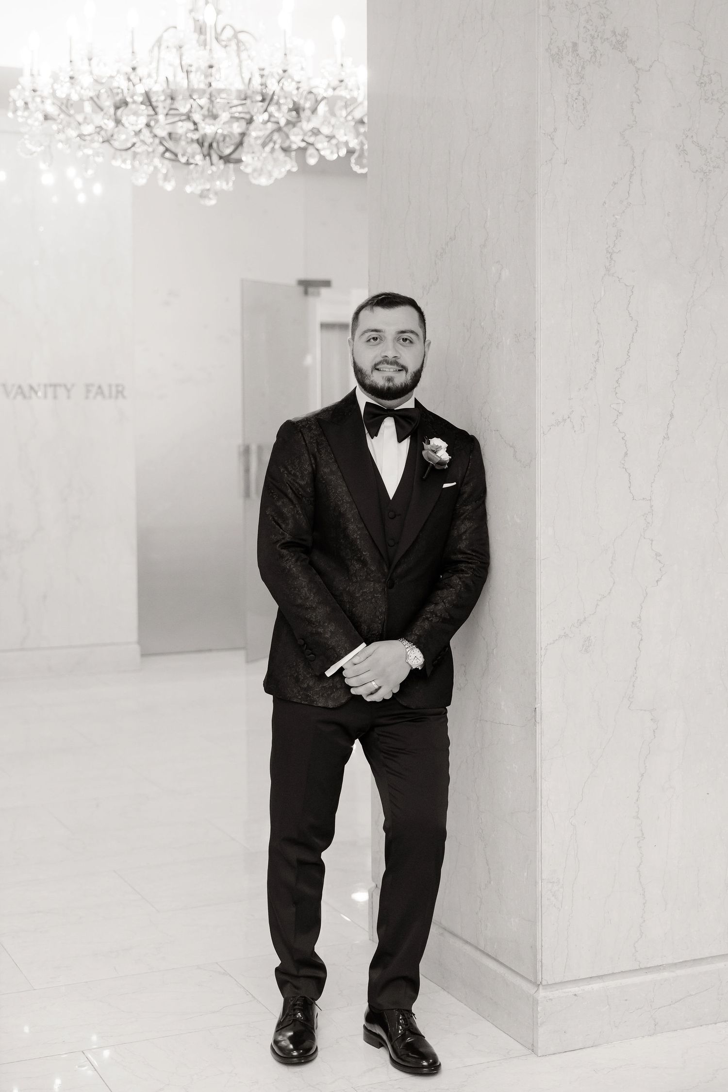 Black and white portraits of a groom in formal attire against a marble wall backdrop.