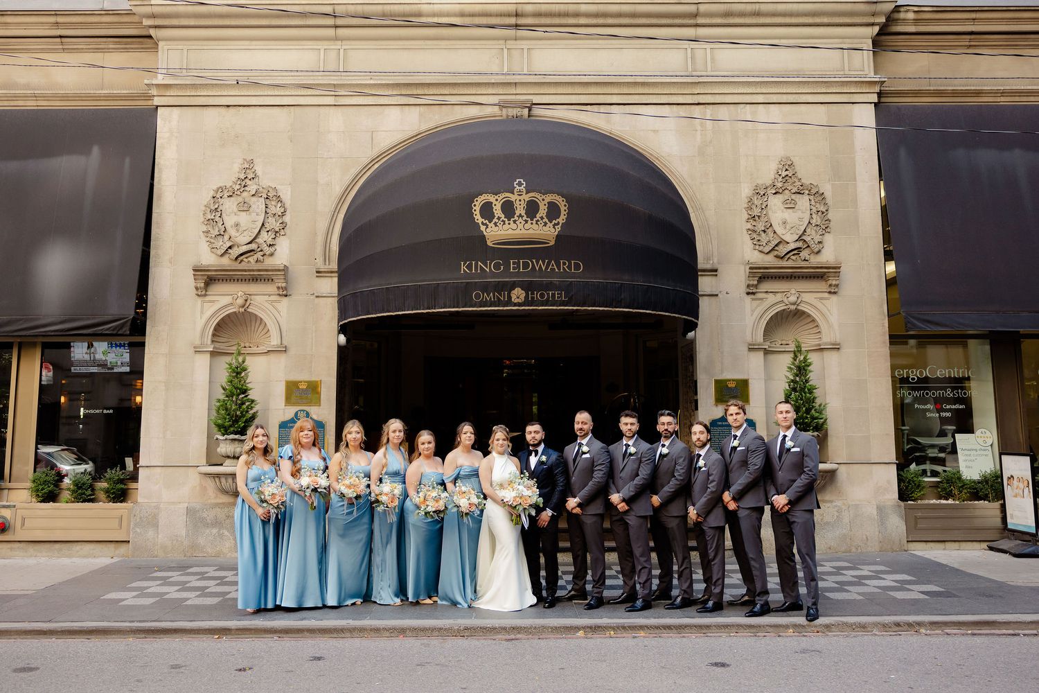 Wedding party in light blue and black attire poses outside historic stone building with crown signage.