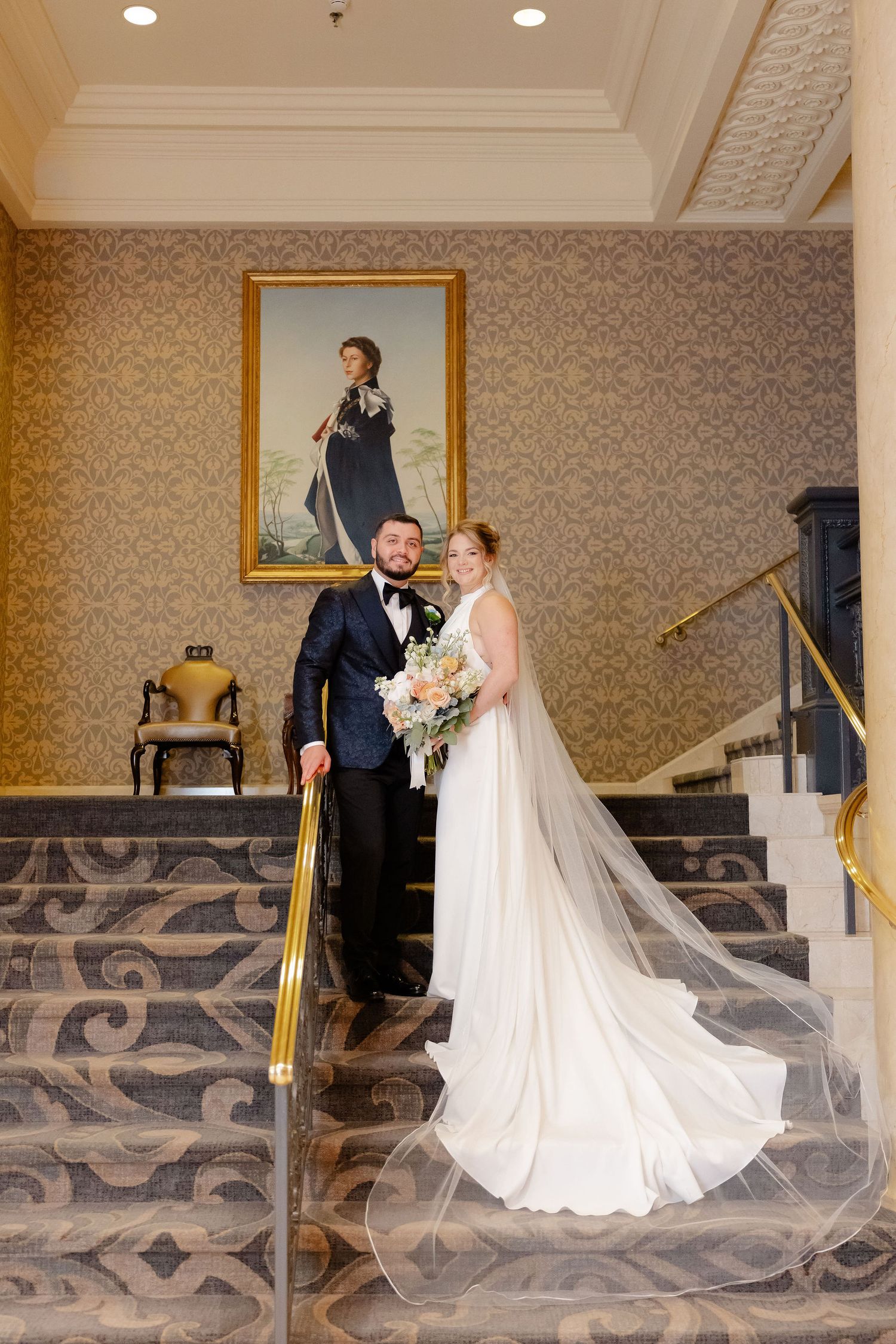 A couple descends an elegant staircase with gold railings and patterned carpeting during their wedding.