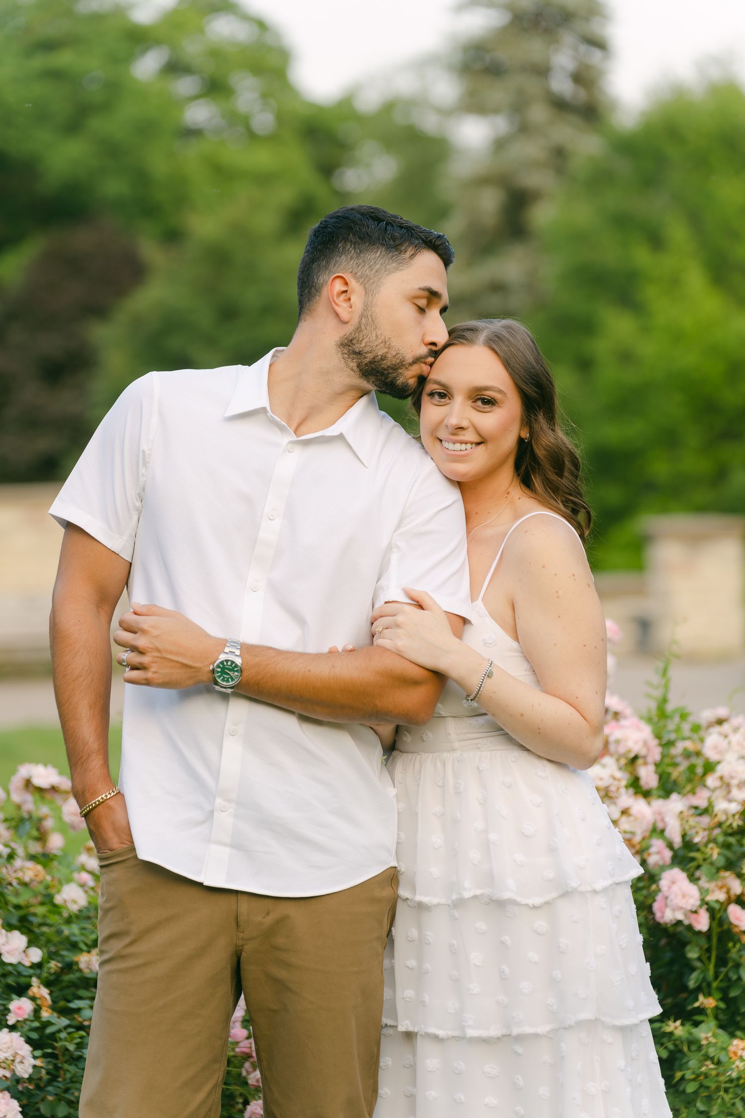 Couple embraces in a romantic garden setting surrounded by pink flowers and greenery.