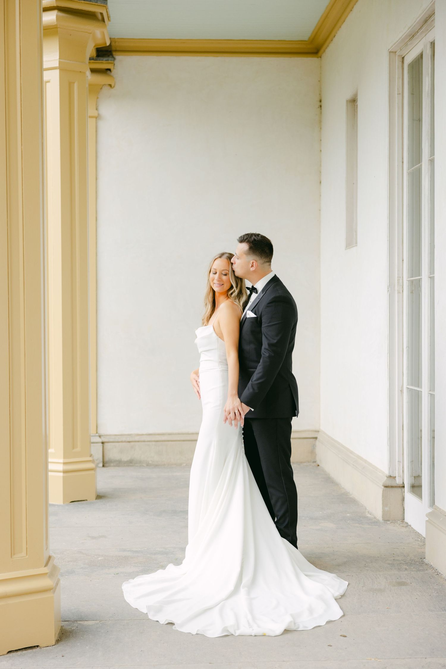 Wedding couple standing in romantic pose under ornate portico with gold trim.