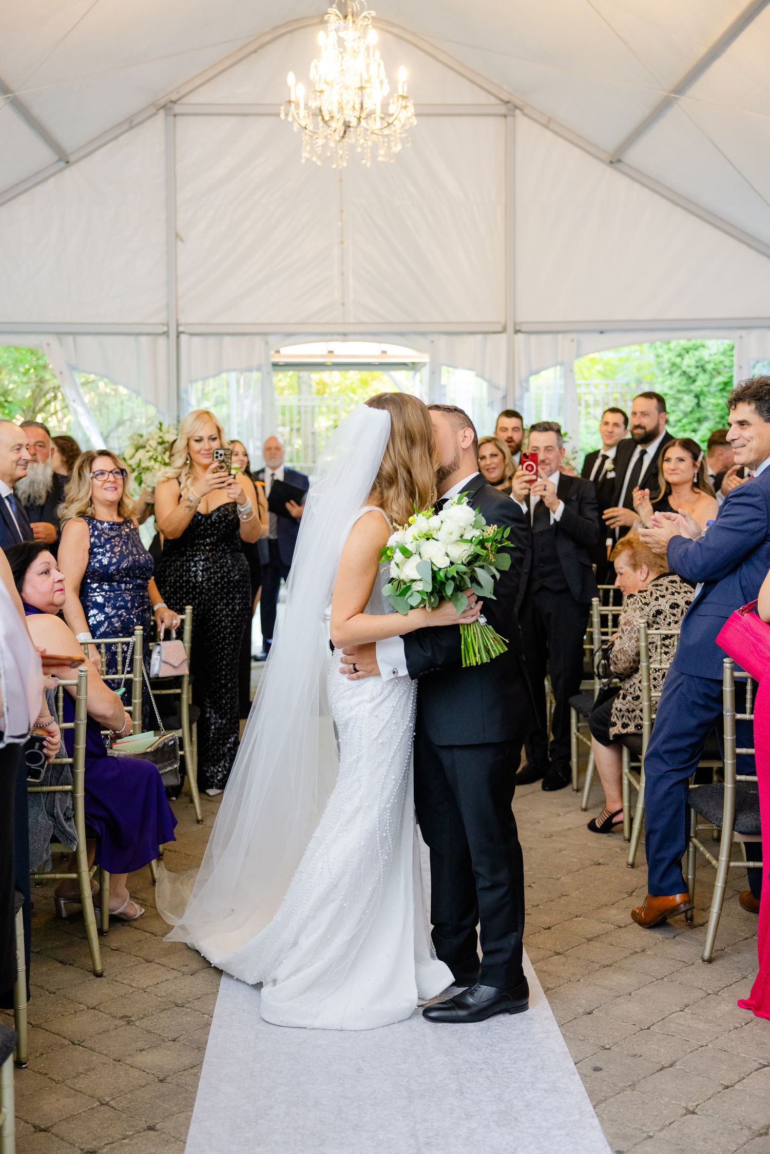 Wedding ceremony moment in an elegantly decorated white tent with a crystal chandelier and seated guests.