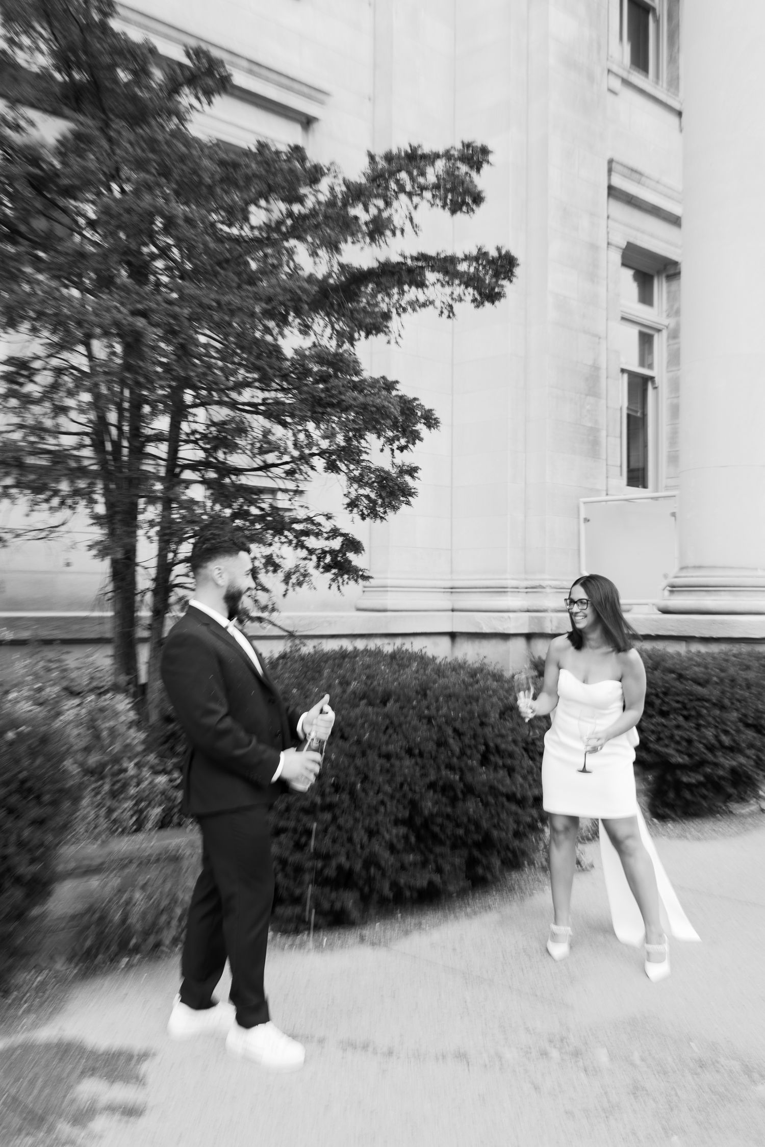 A black and white photo of a couple sharing champagne outside an elegant building.