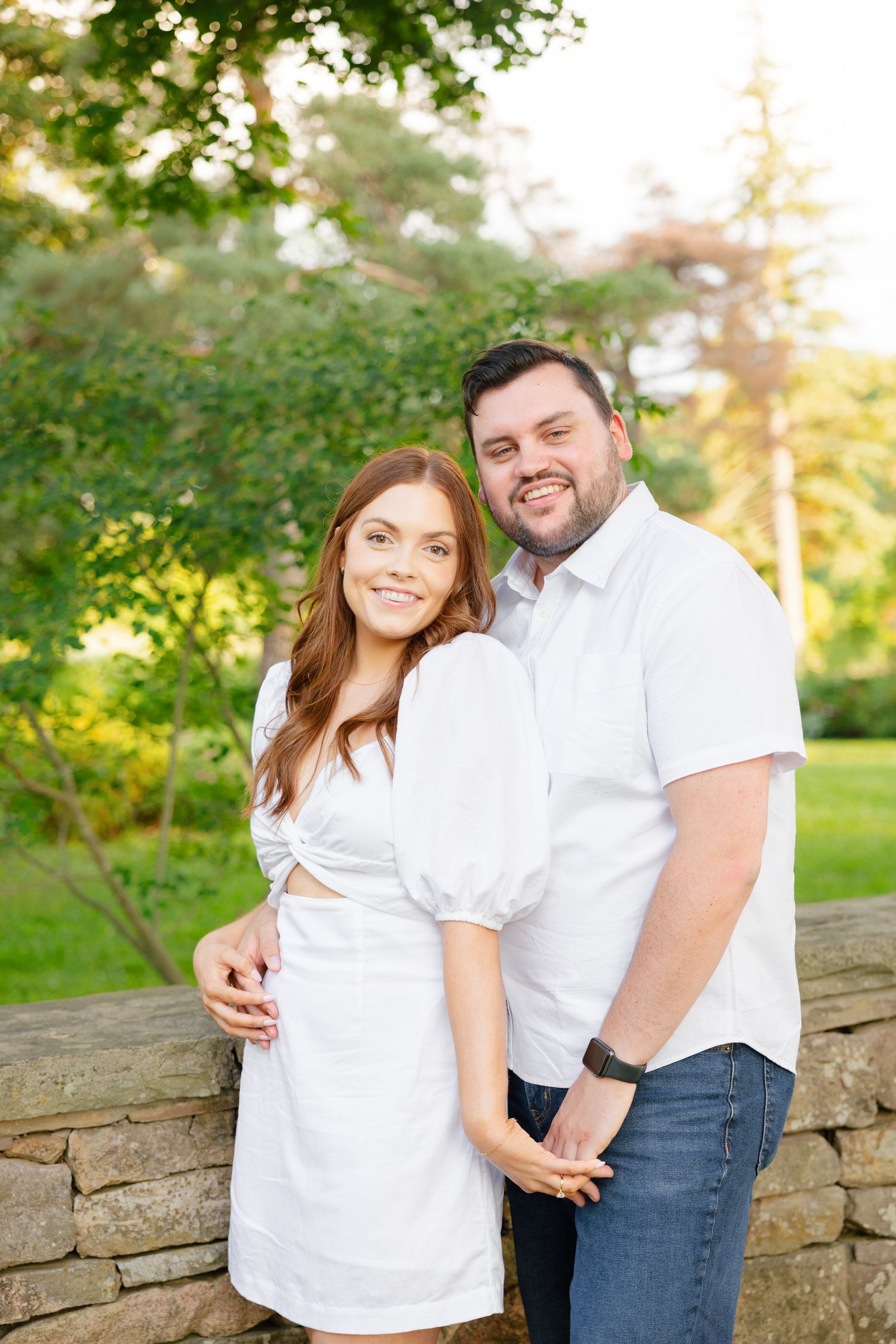 A happy couple in matching white outfits poses together against a stone wall outdoors.
