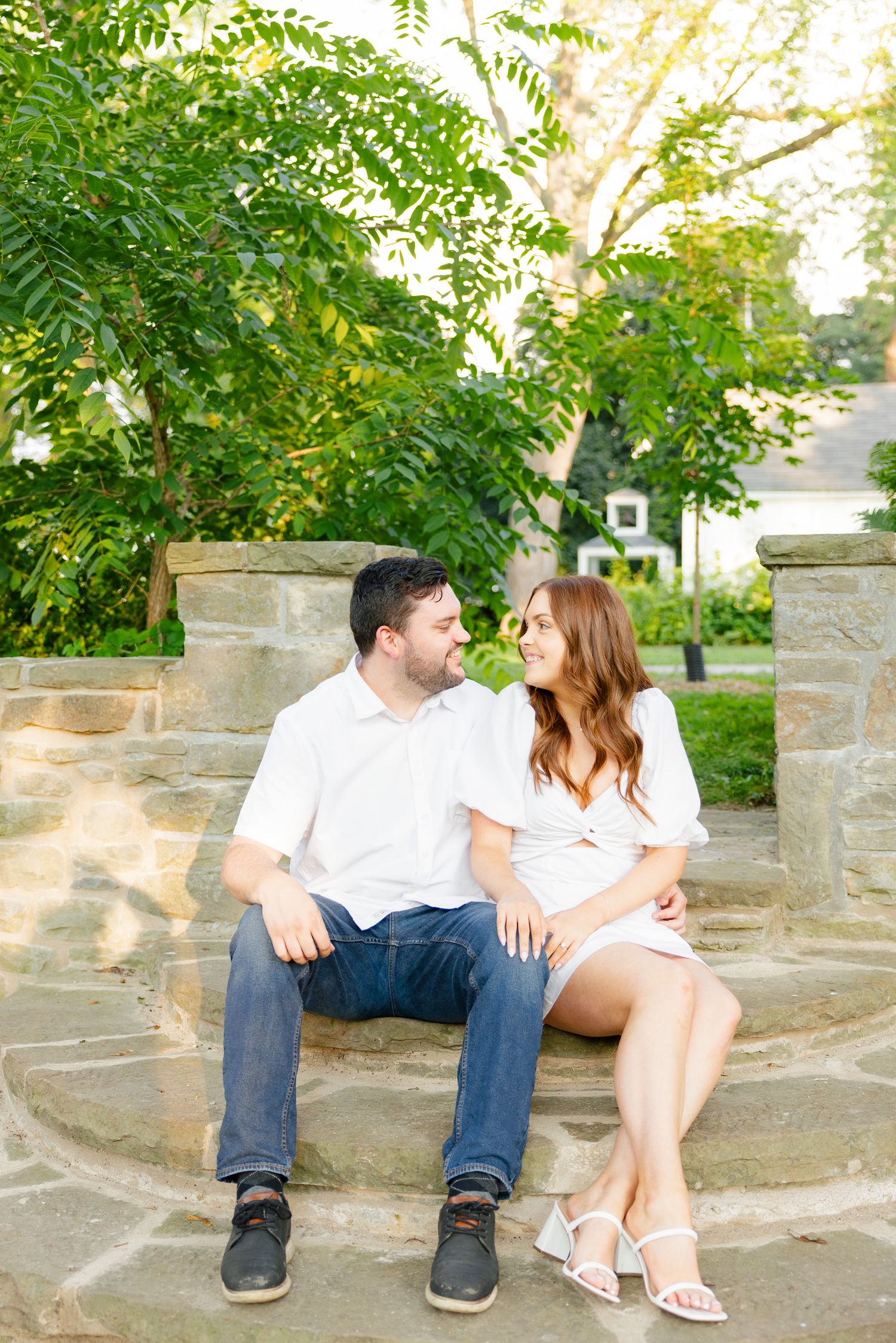 Couple in coordinating white outfits sitting together on stone steps surrounded by green foliage.