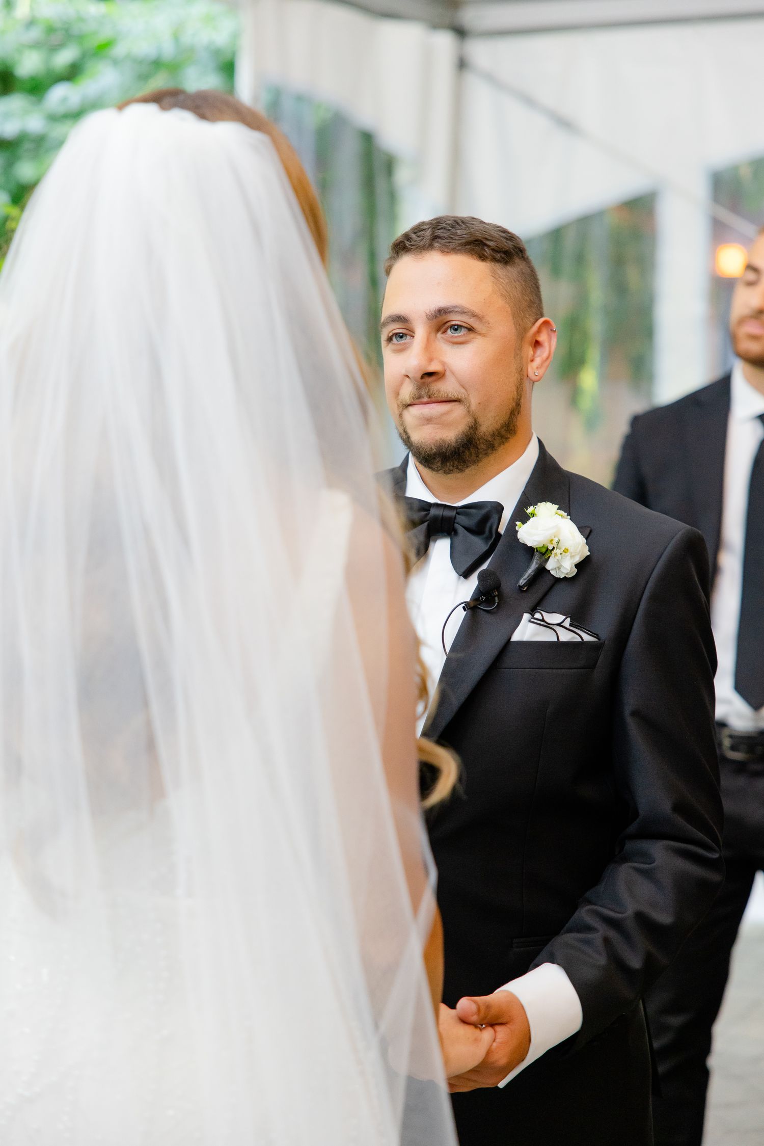 Wedding ceremony moment in outdoor tent with groom in black tuxedo and bow tie.