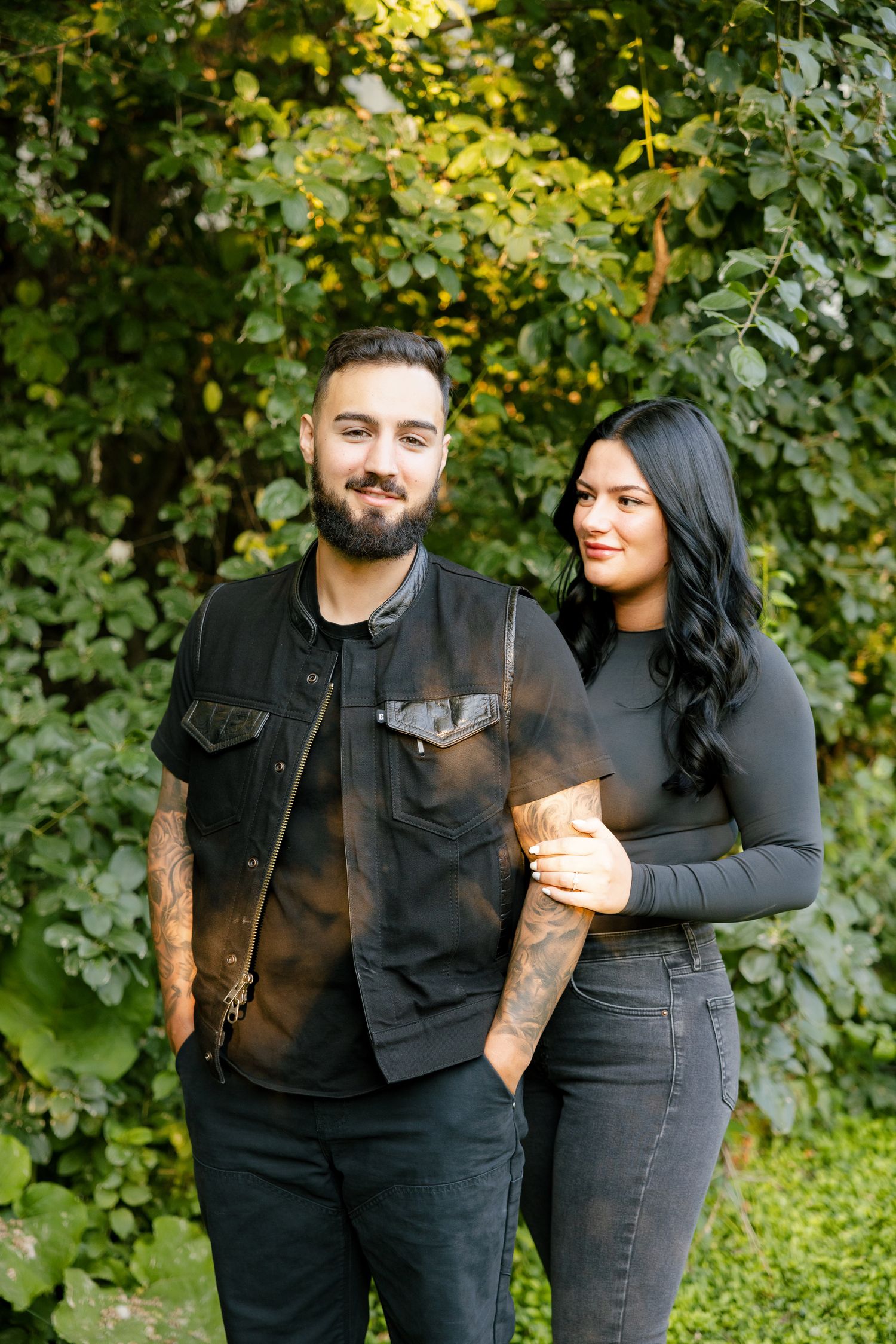 A couple in dark casual wear poses together against a background of summer foliage.