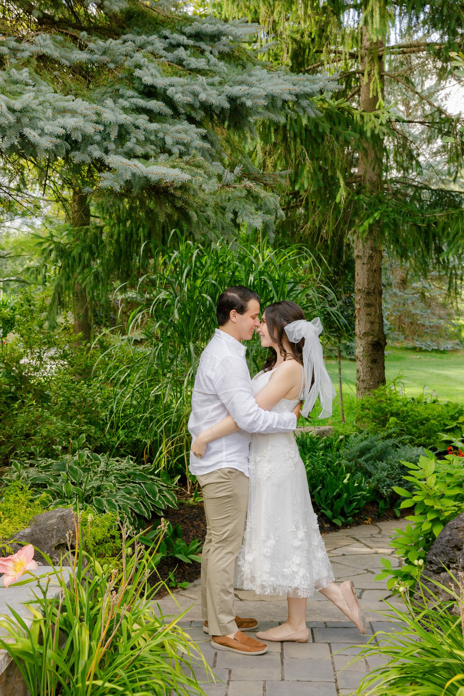 Romantic wedding moment captured on garden path beneath towering evergreen trees.