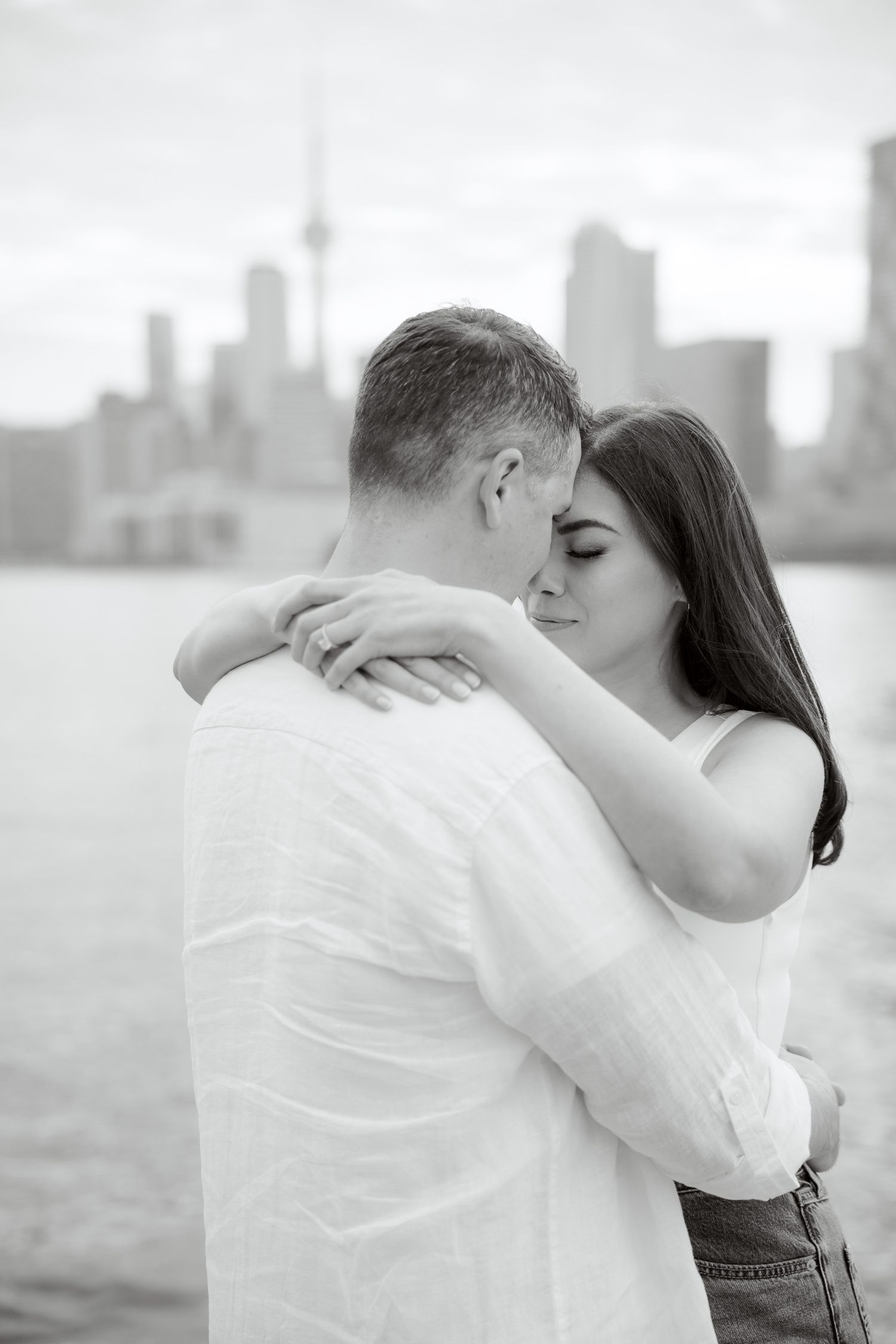 Romantic couple embraces against Toronto skyline backdrop in black and white photograph.