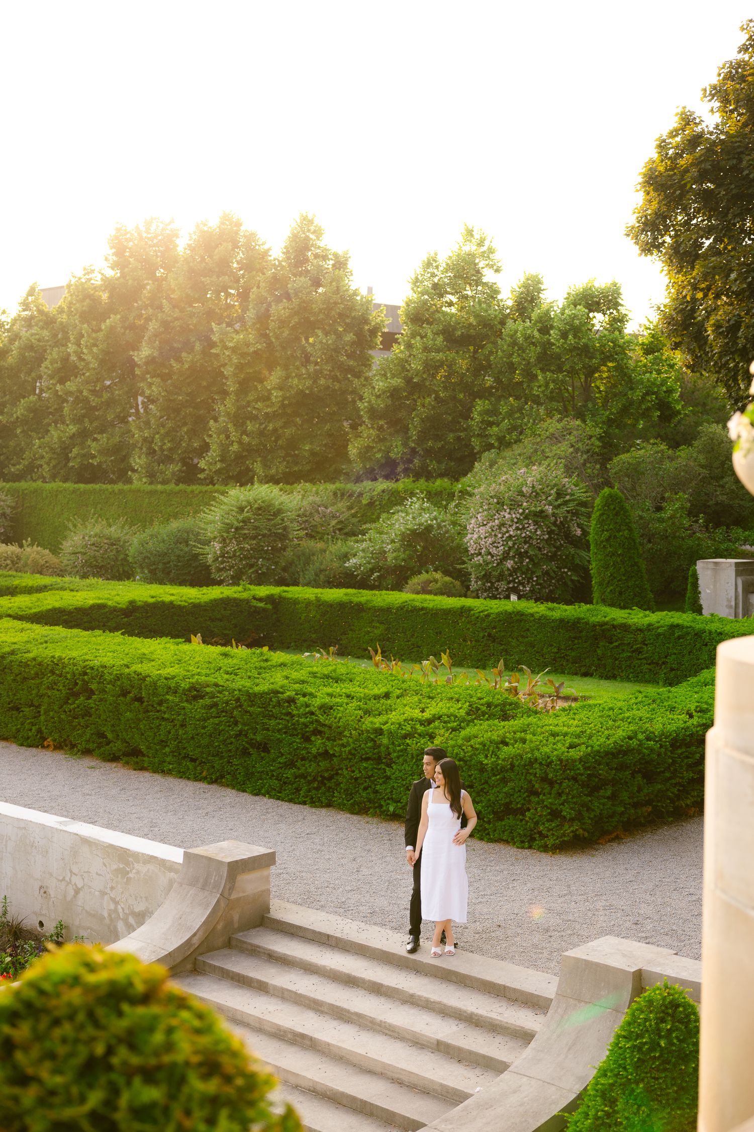 A person in a white dress standing on elegant stone steps surrounded by manicured gardens at sunset.