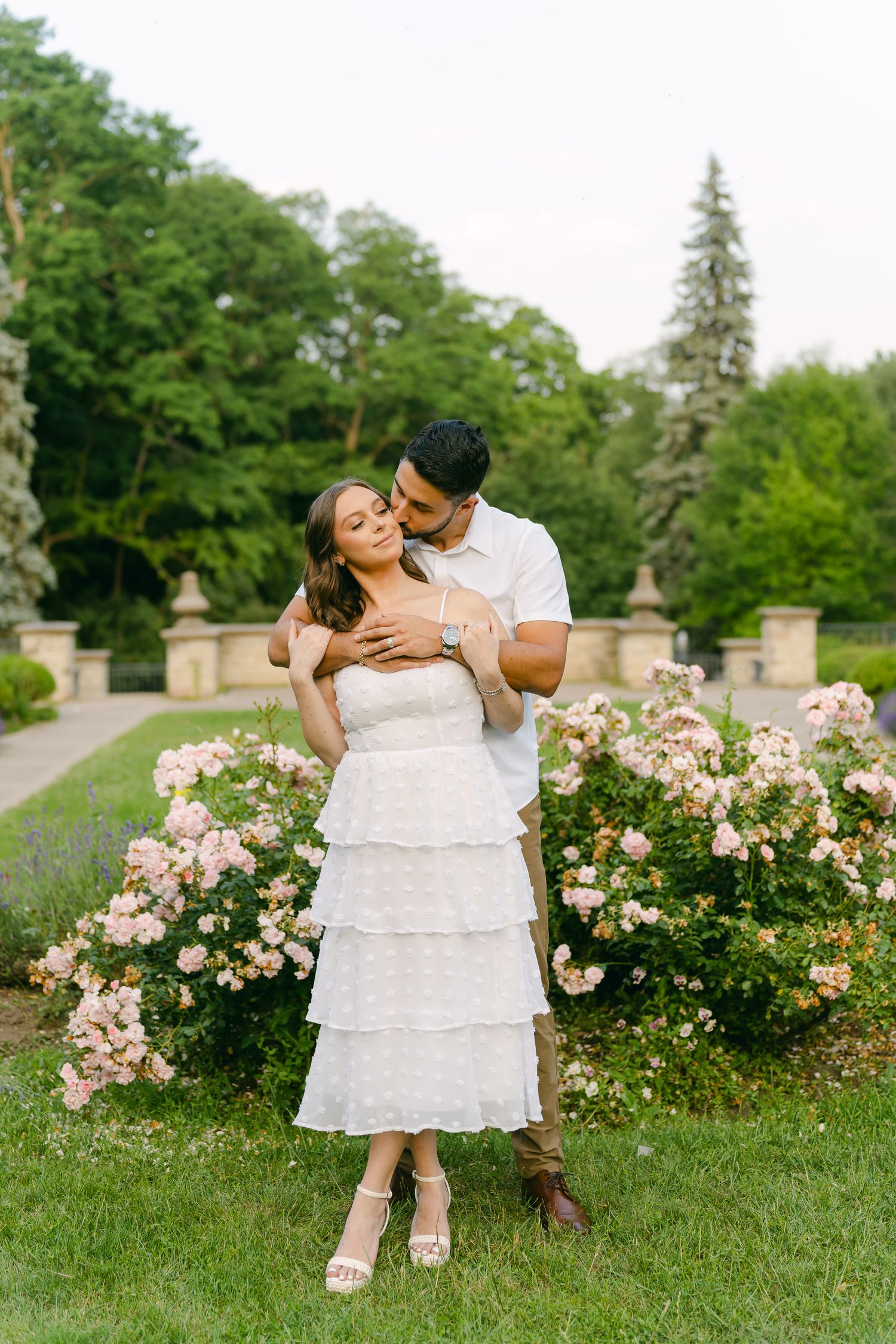 A romantic couple embraces in a garden setting with blooming roses and stone pillars in the background.