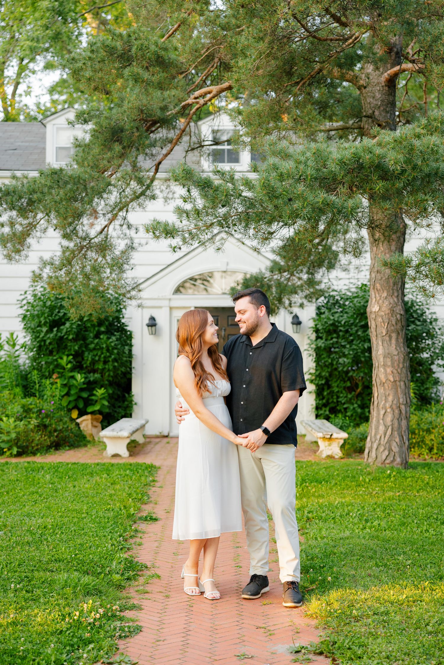 A romantic couple stands together on a brick path in front of a white house with large pine trees.