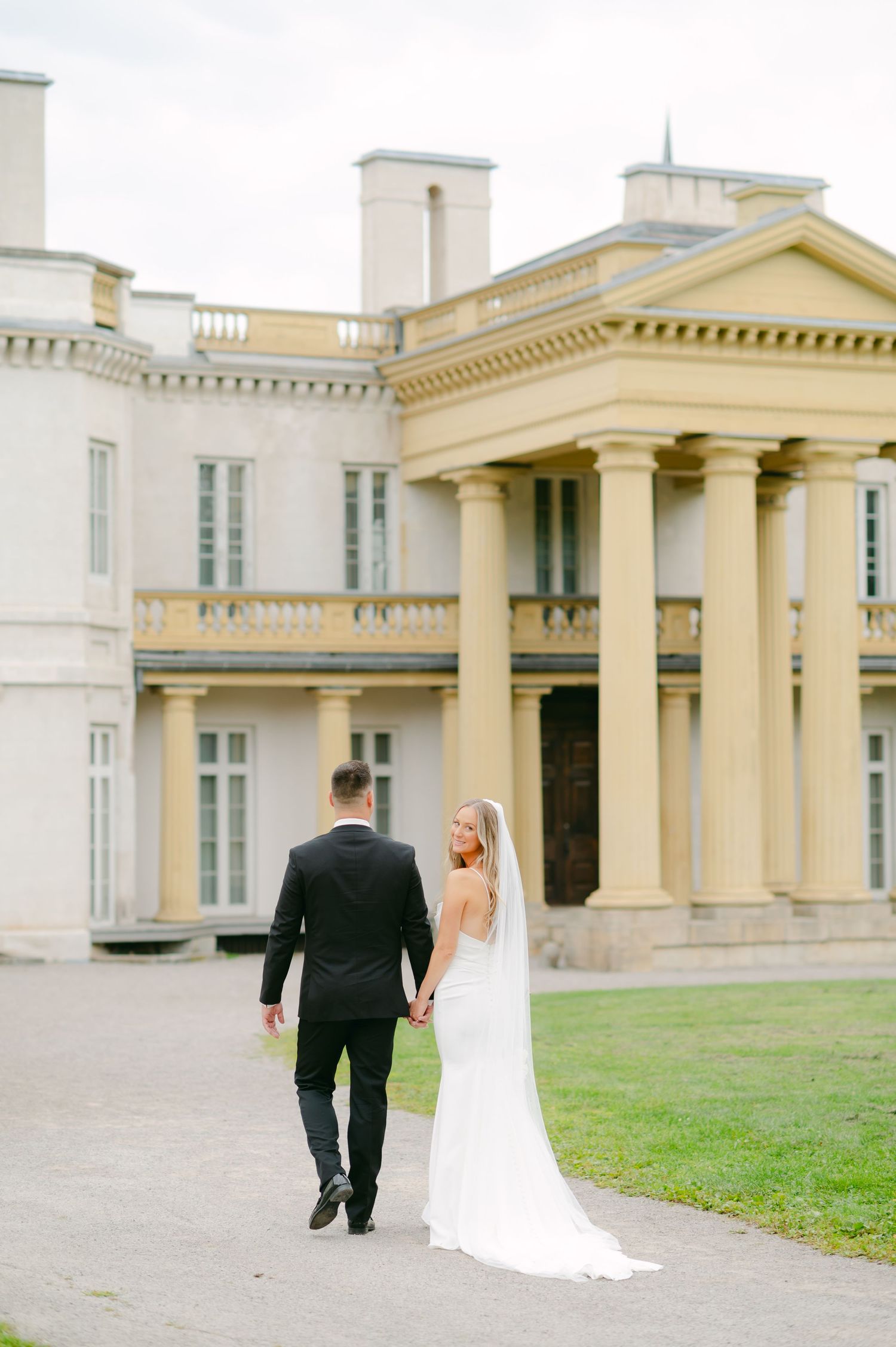 A couple in wedding attire walking along the path of a stately historic building.
