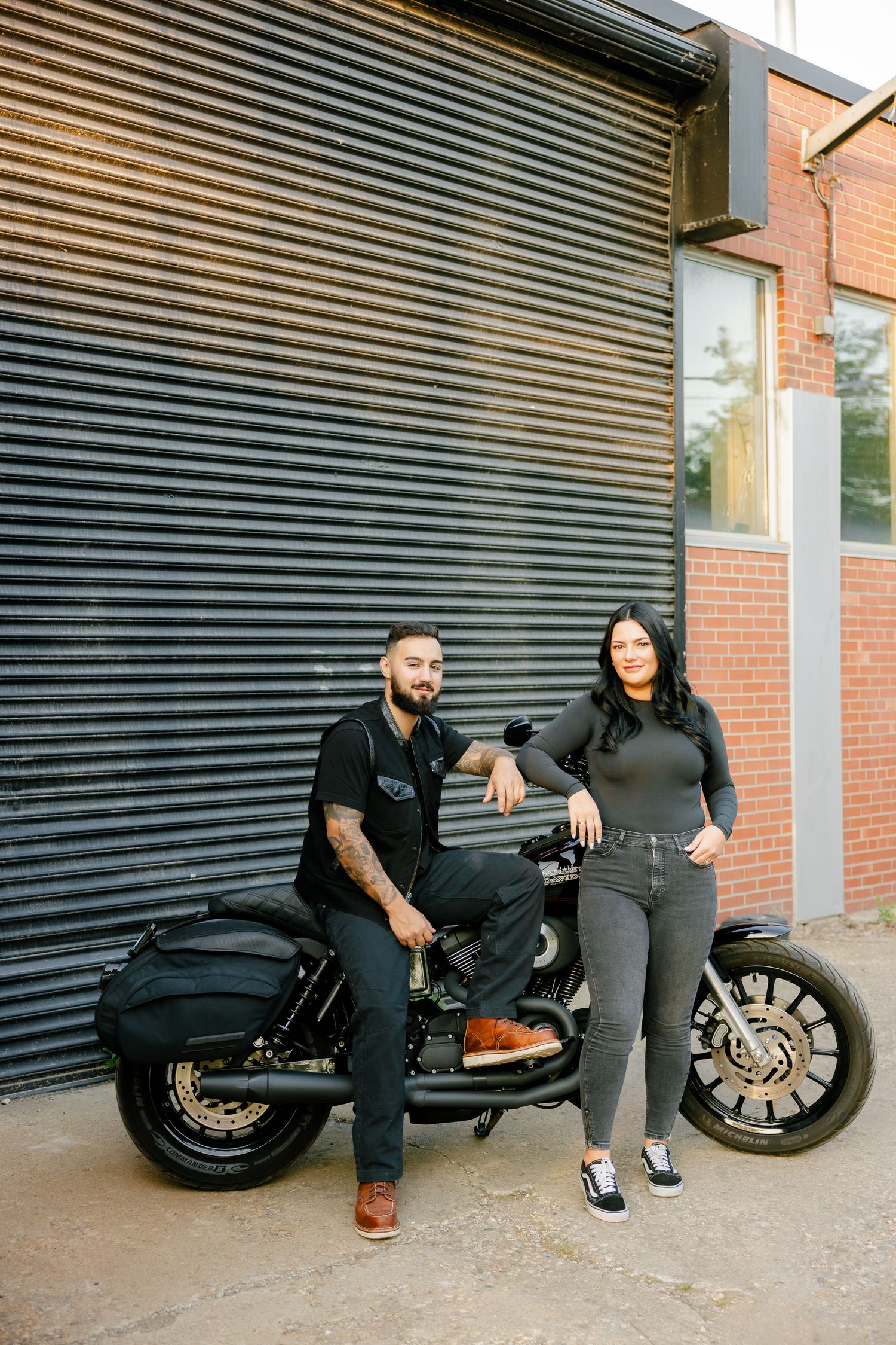 Casual portrait of two people with their dark motorcycle against industrial architecture.