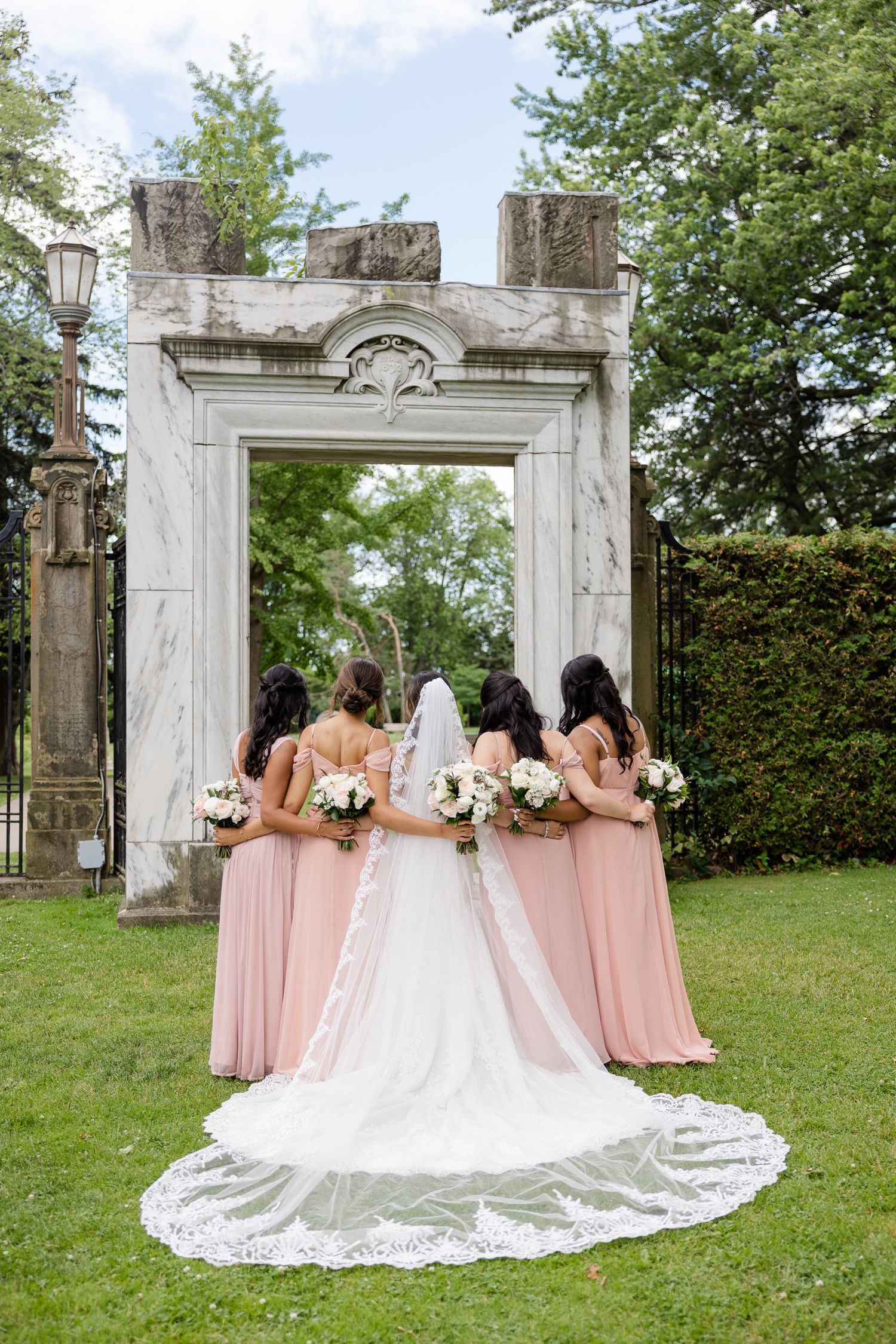 Bridesmaids in blush pink dresses stand behind bride with cathedral train at ornate stone archway.