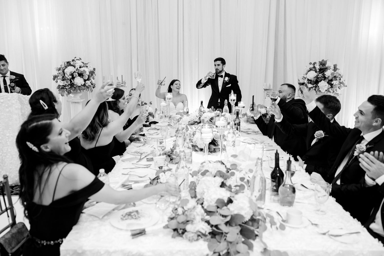 Black and white photo of wedding reception guests toasting at an elegant dinner table.