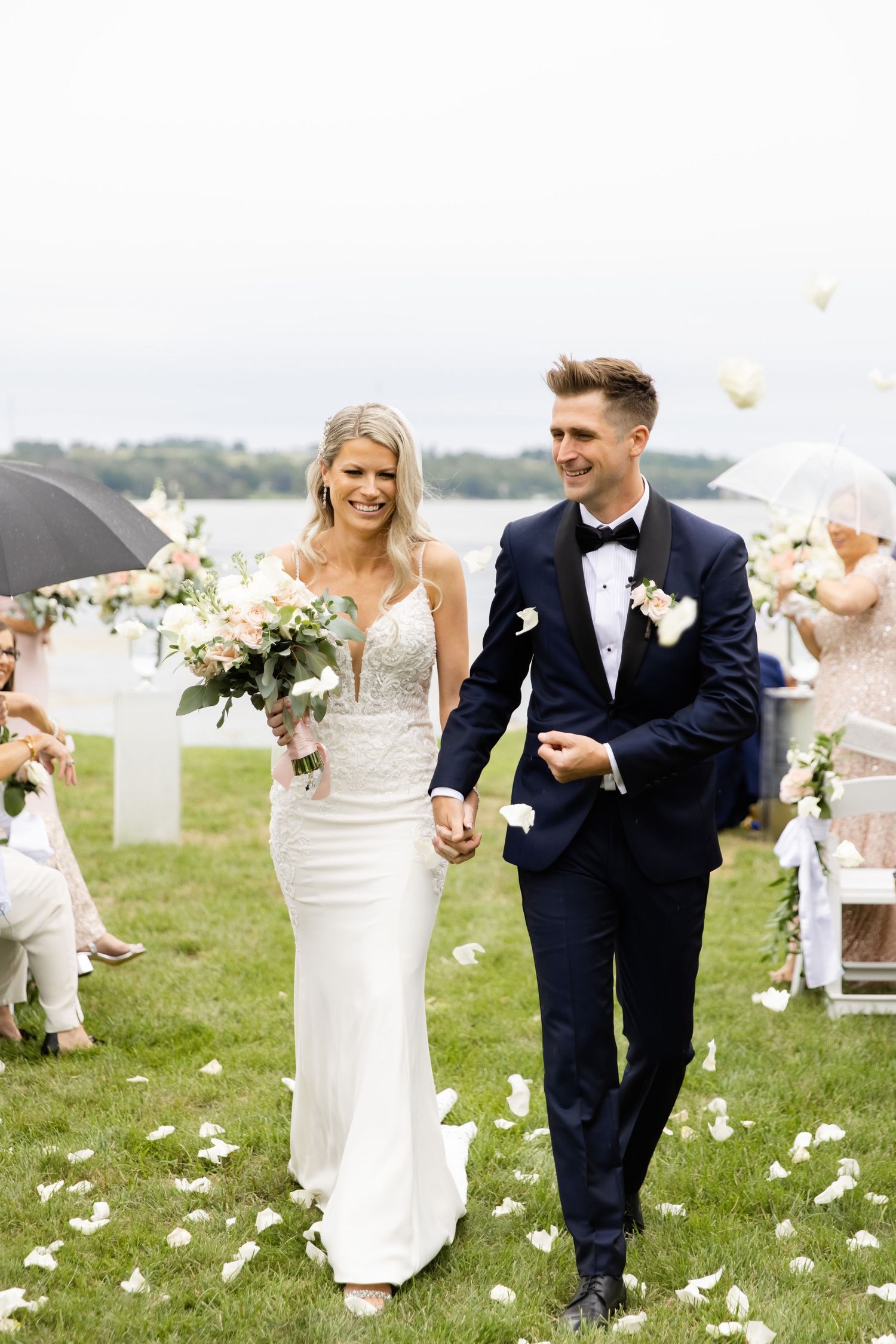 Happy couple exits ceremony under shower of white petals on grassy lawn.