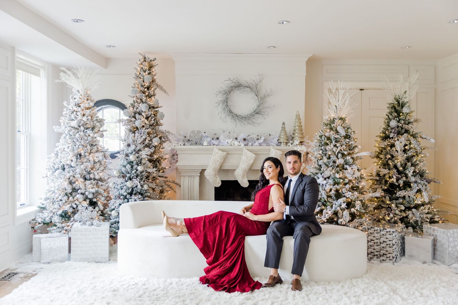 Festive portrait session in bright white room with multiple decorated Christmas trees and red gown.