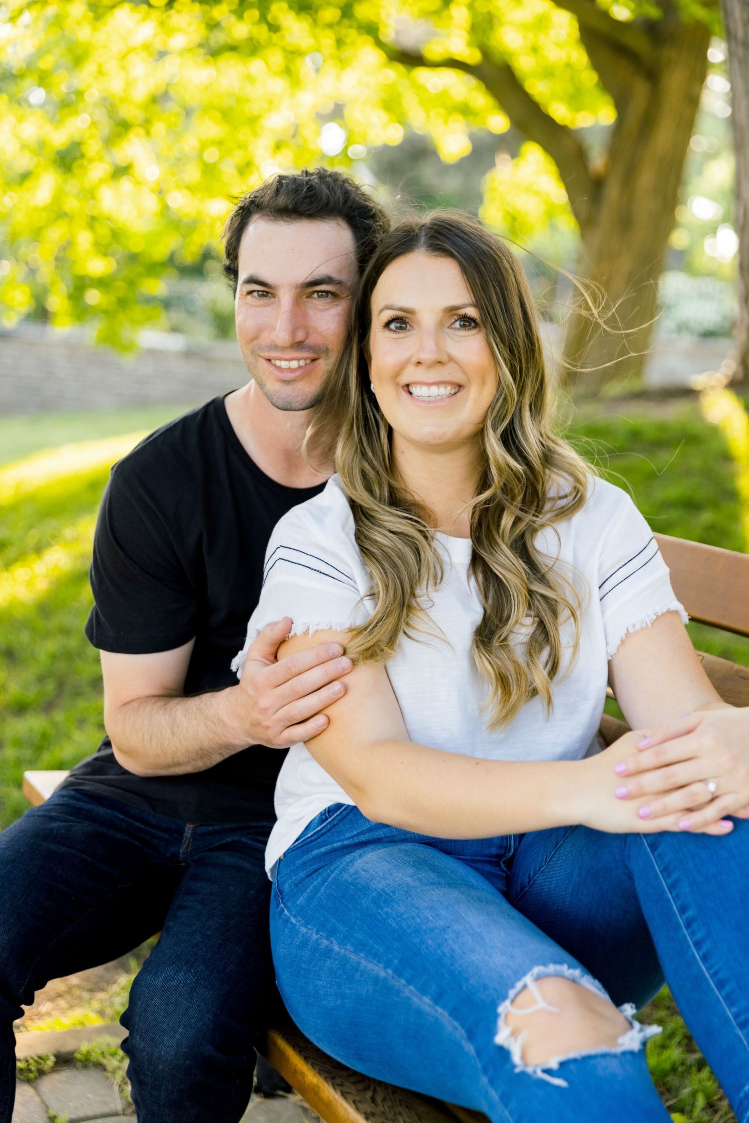 A couple sits together on a bench surrounded by sunlit trees in a park setting.