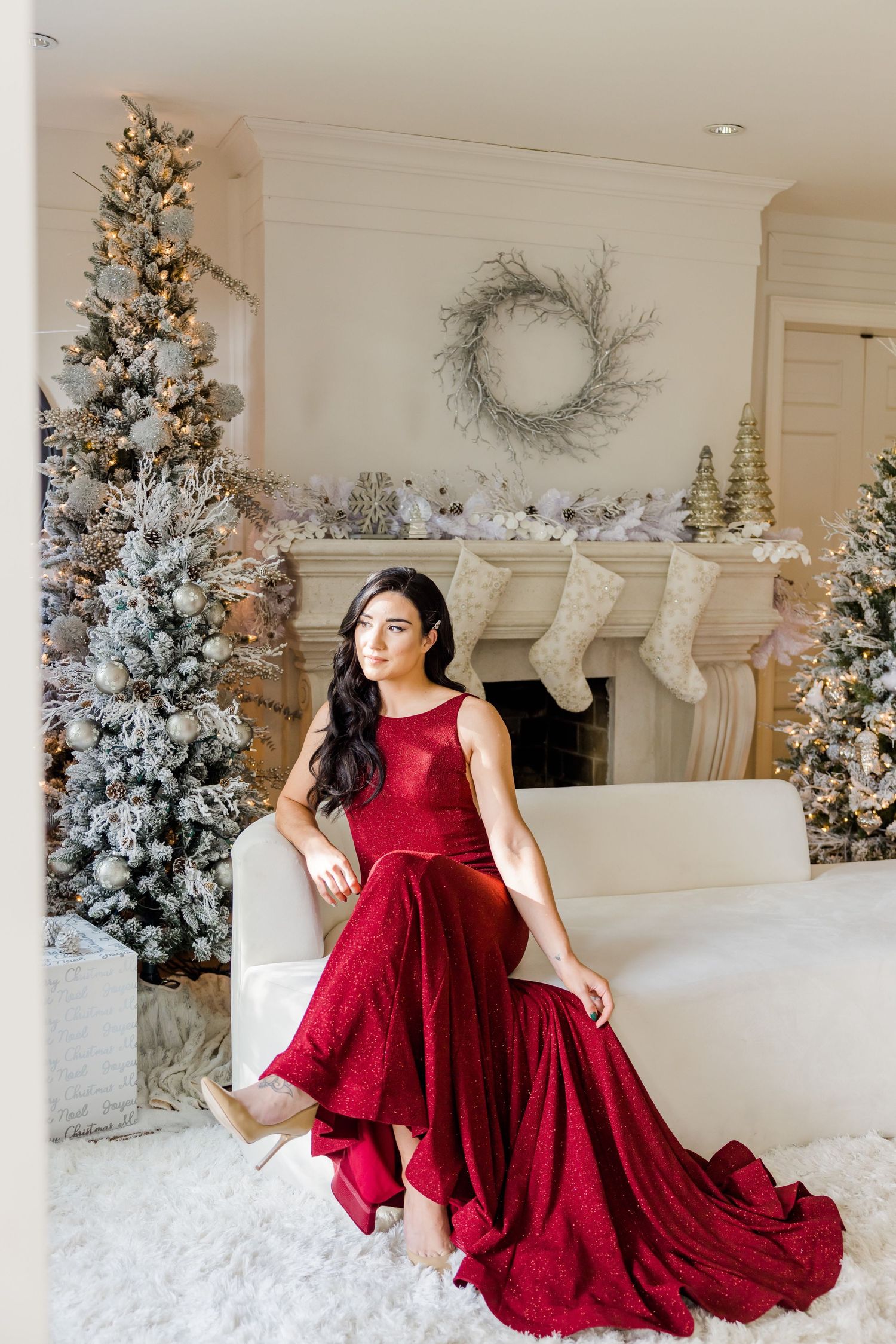 Red velvet gown photographed against elegant white Christmas decor with flocked trees and stockings on fireplace mantel.