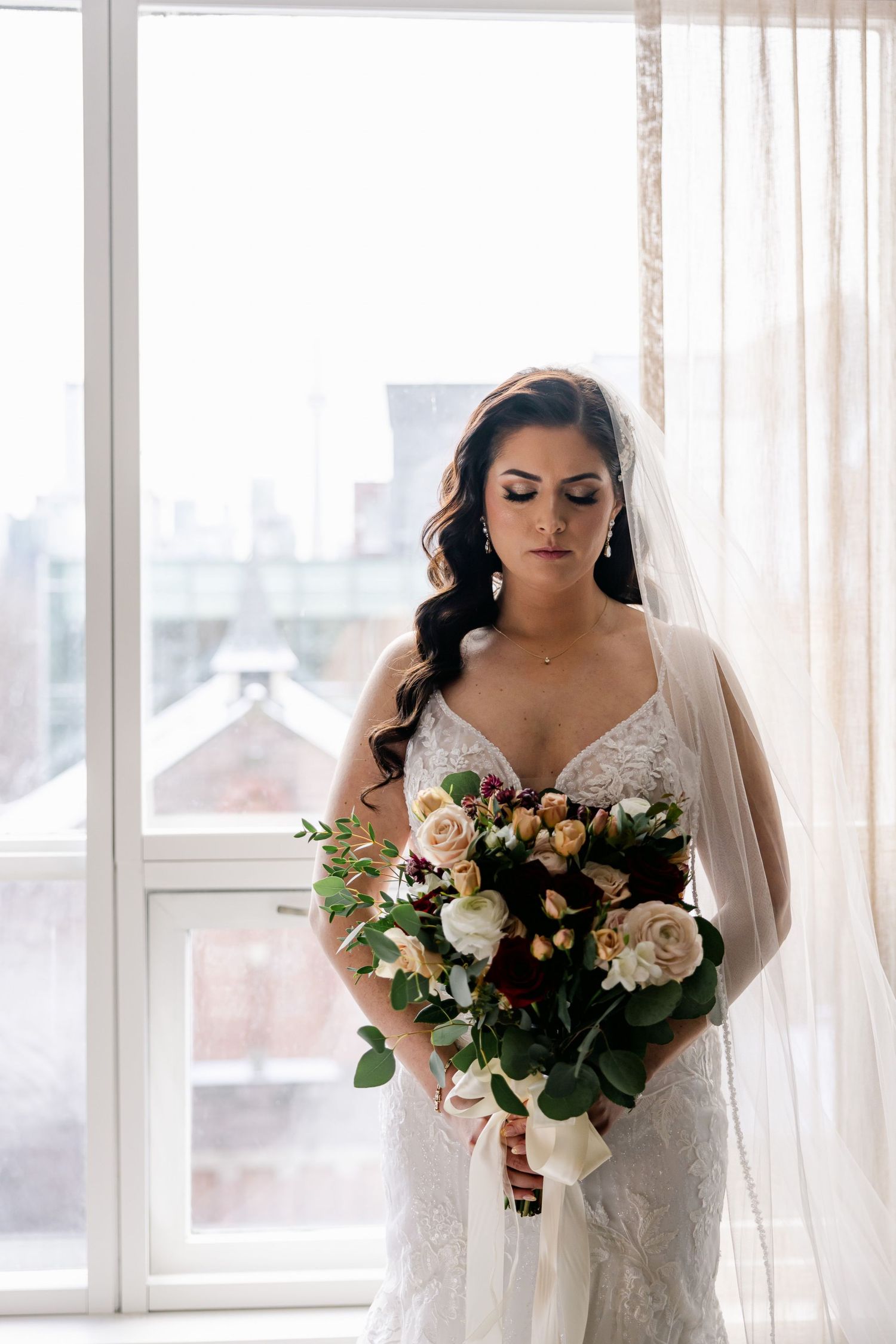 A bride holds a lush bouquet of roses and greenery by a sunlit window.
