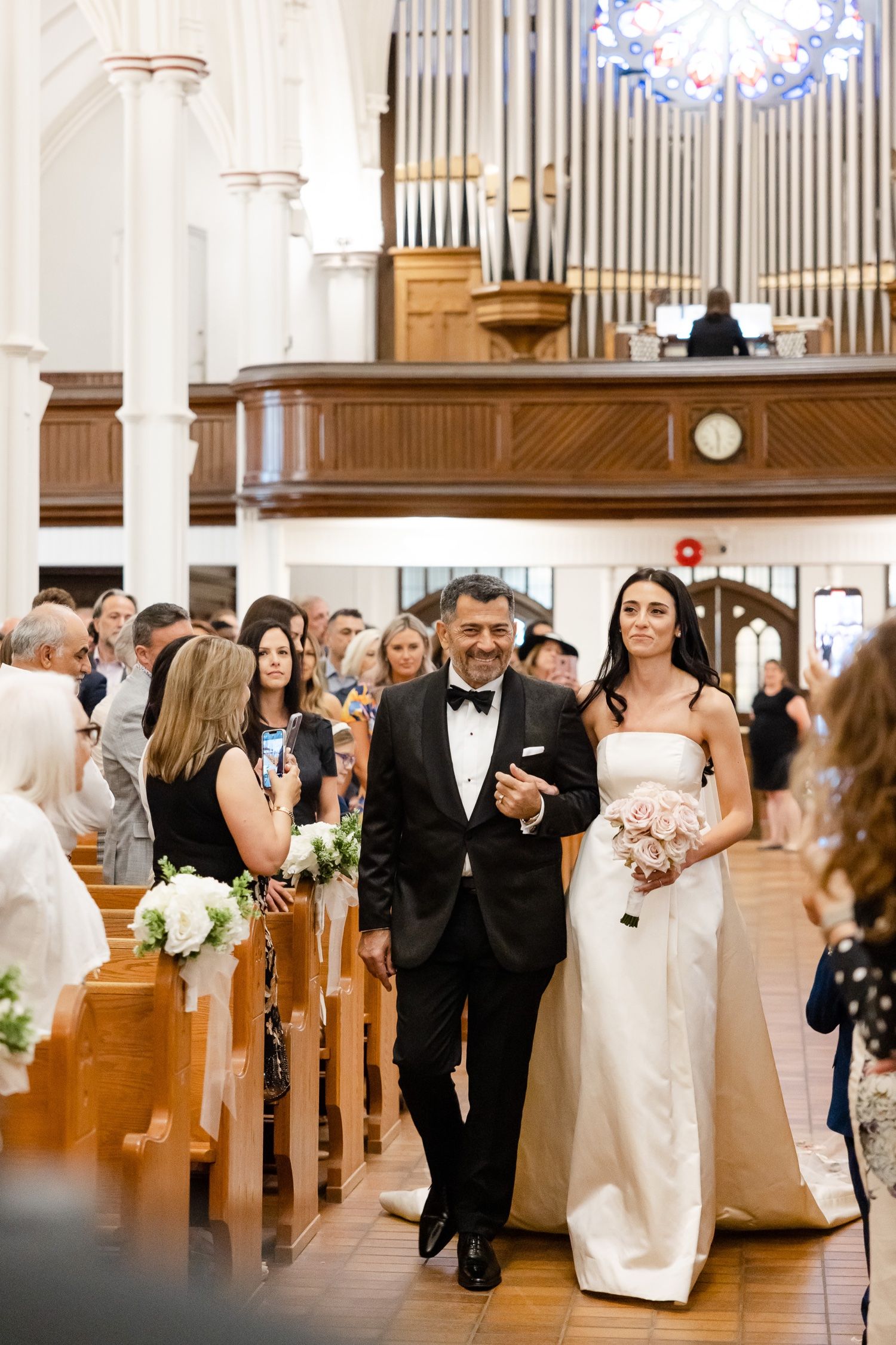 Wedding party walking down the church aisle as guests look on with organ pipes visible in background.