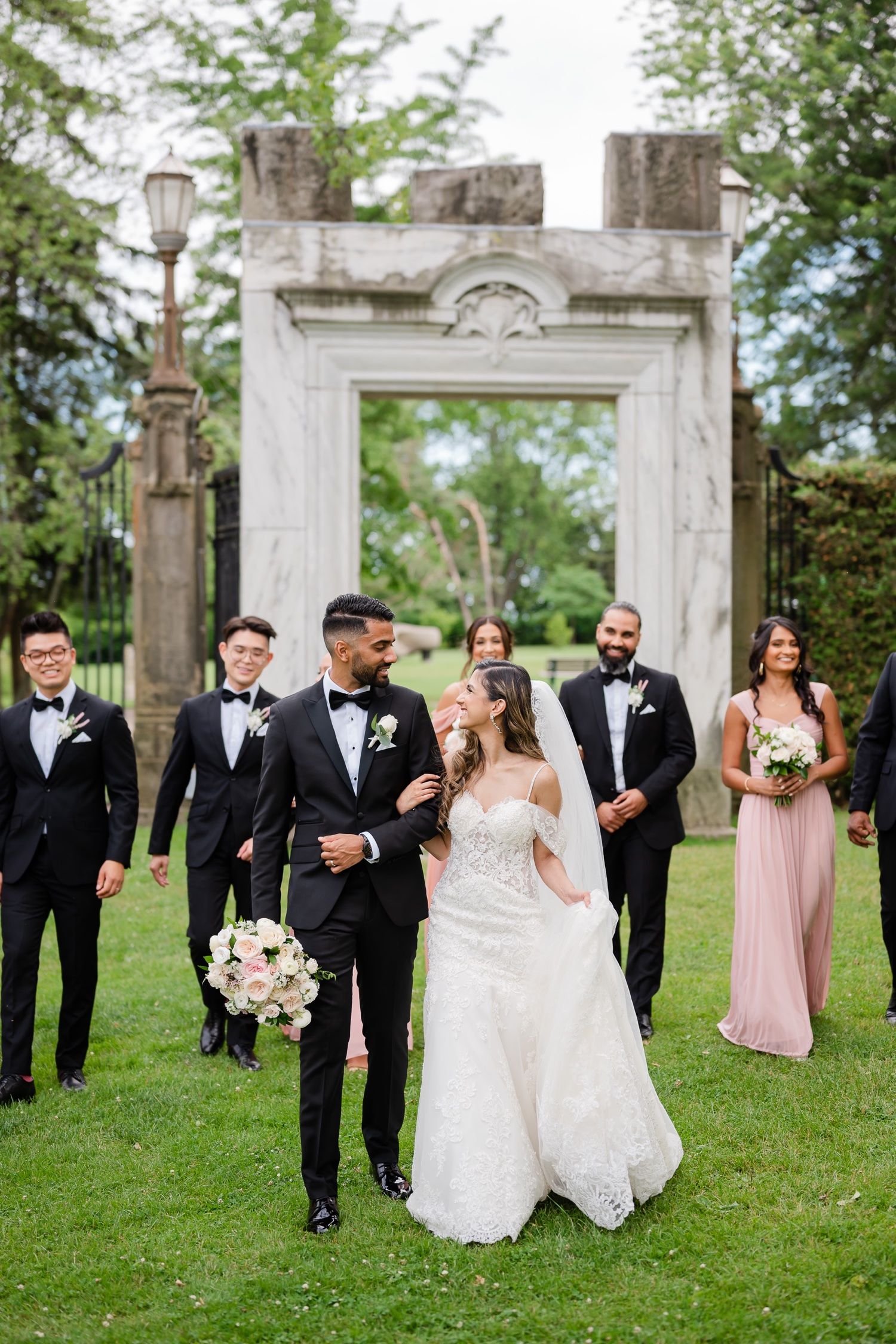 Candid wedding party moment captured in front of a stone archway with vintage lamp posts.