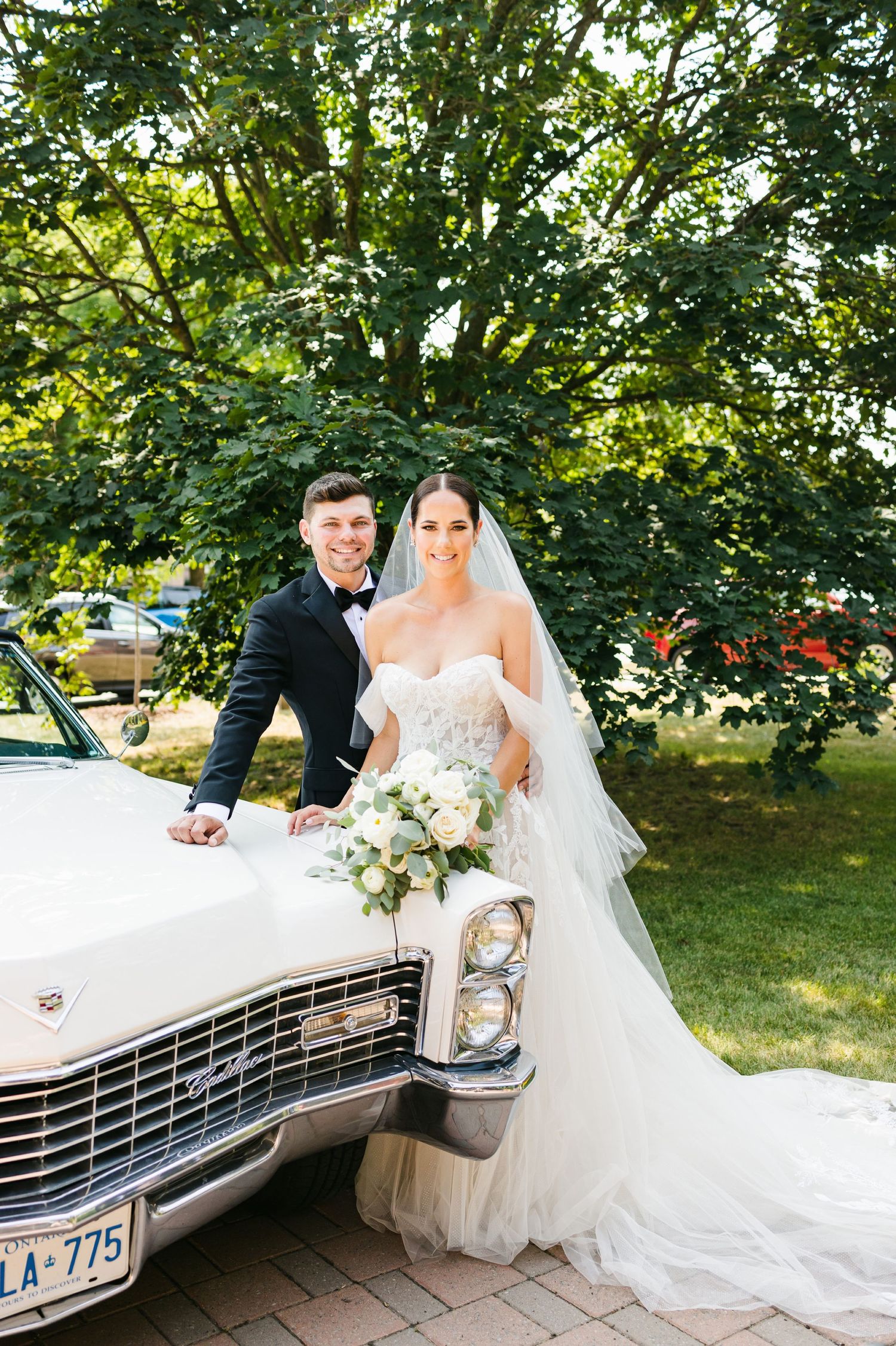 Outdoor wedding portrait with vintage white Cadillac against lush green trees and foliage.
