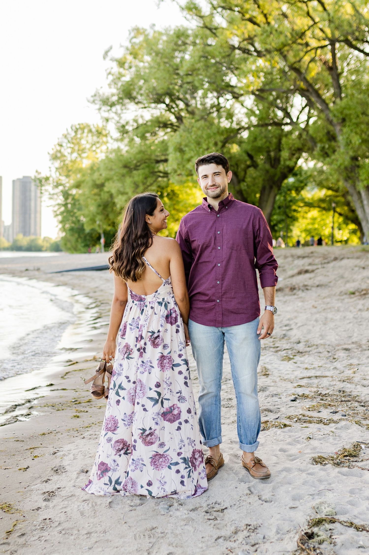 Couple poses together on sandy beach with autumn trees and city skyline.