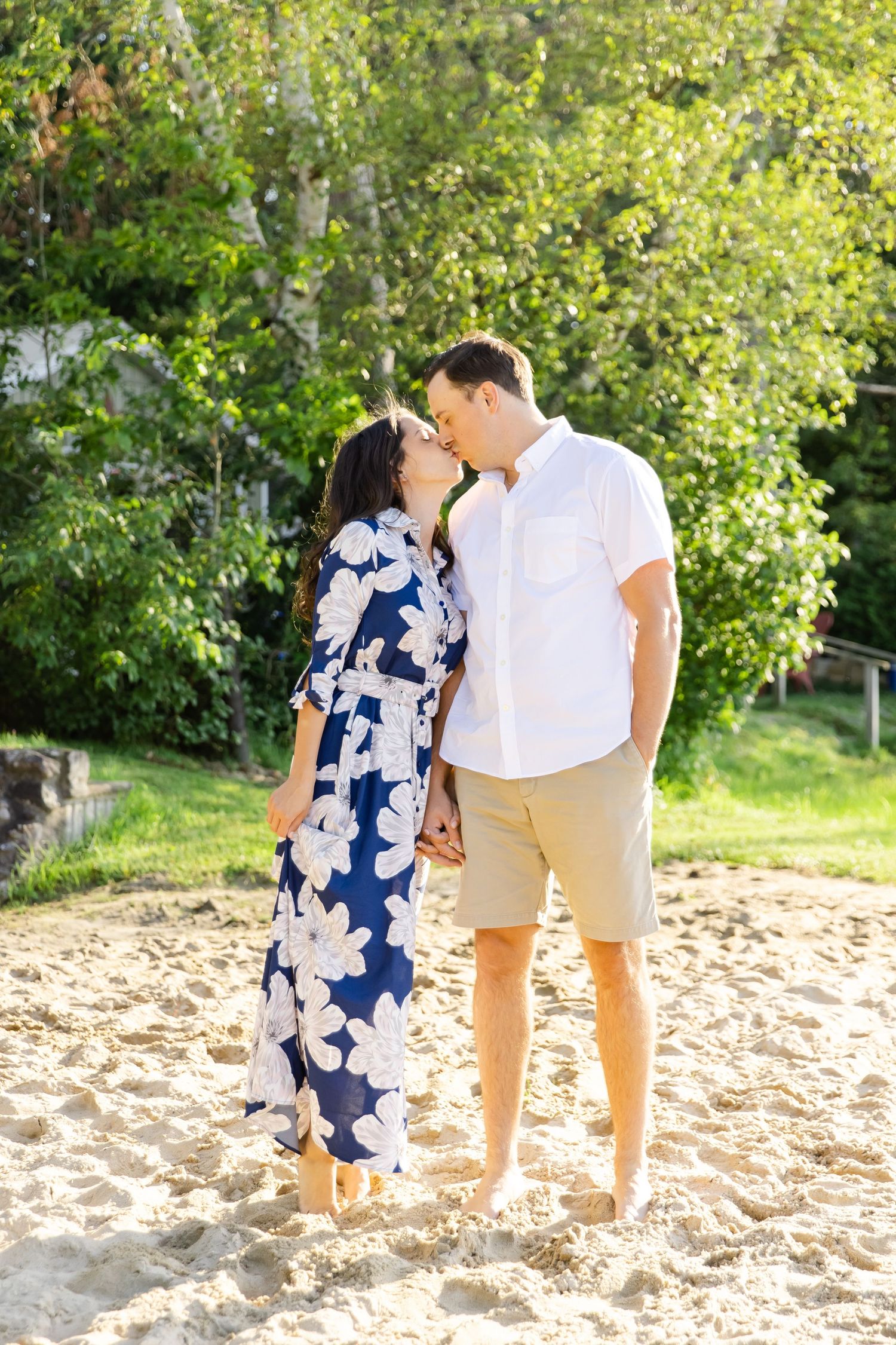 Couple sharing an intimate moment on a sandy beach with lush green foliage in background.