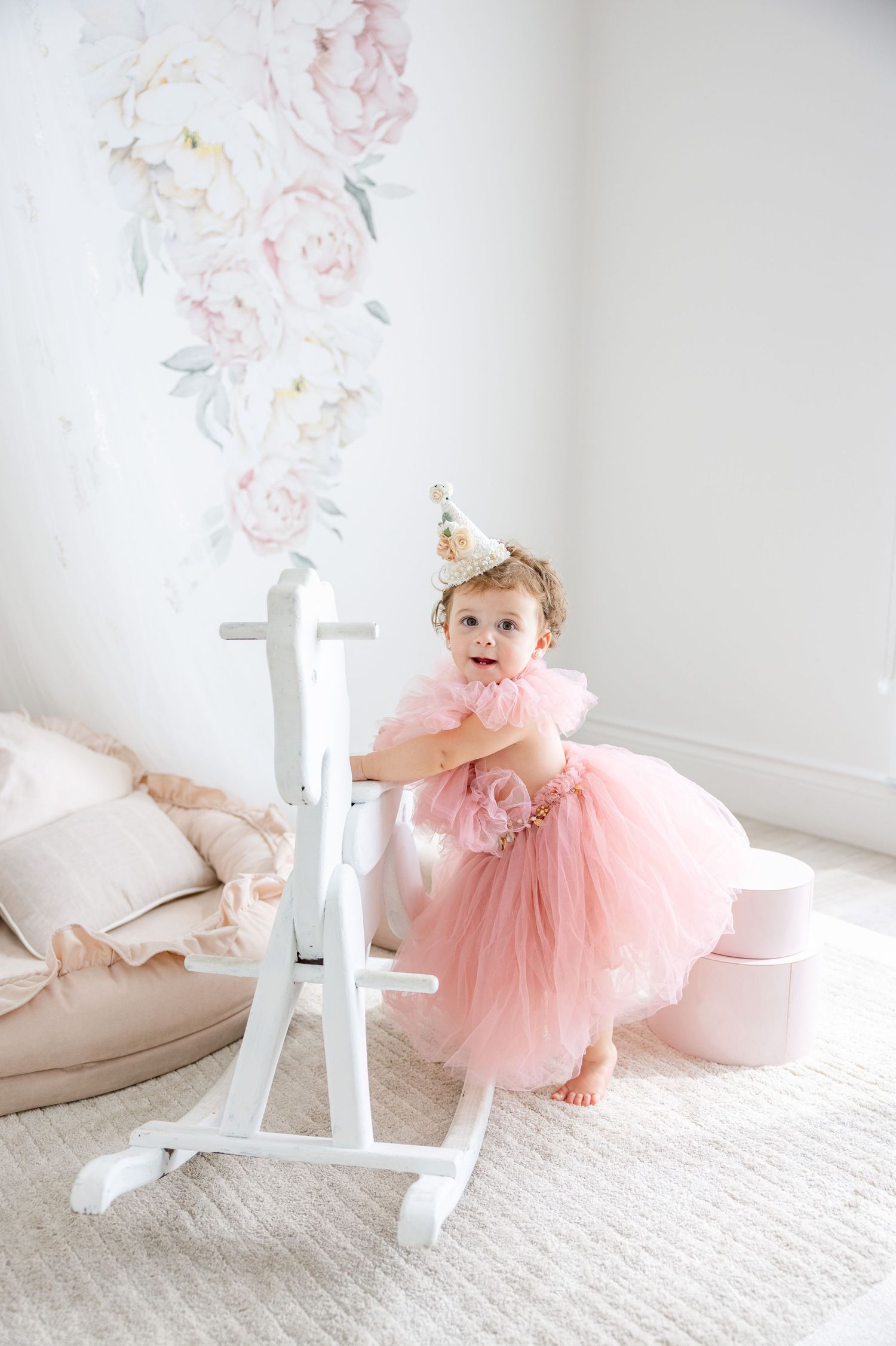 Dreamy nursery setting with white rocking horse and pink party dress against floral backdrop.
