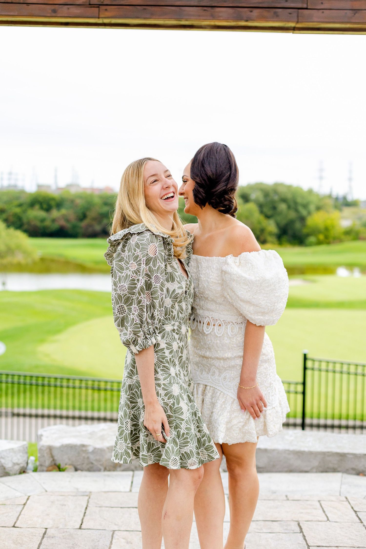 Two friends share a joyful candid moment on stone patio with golf course scenery.