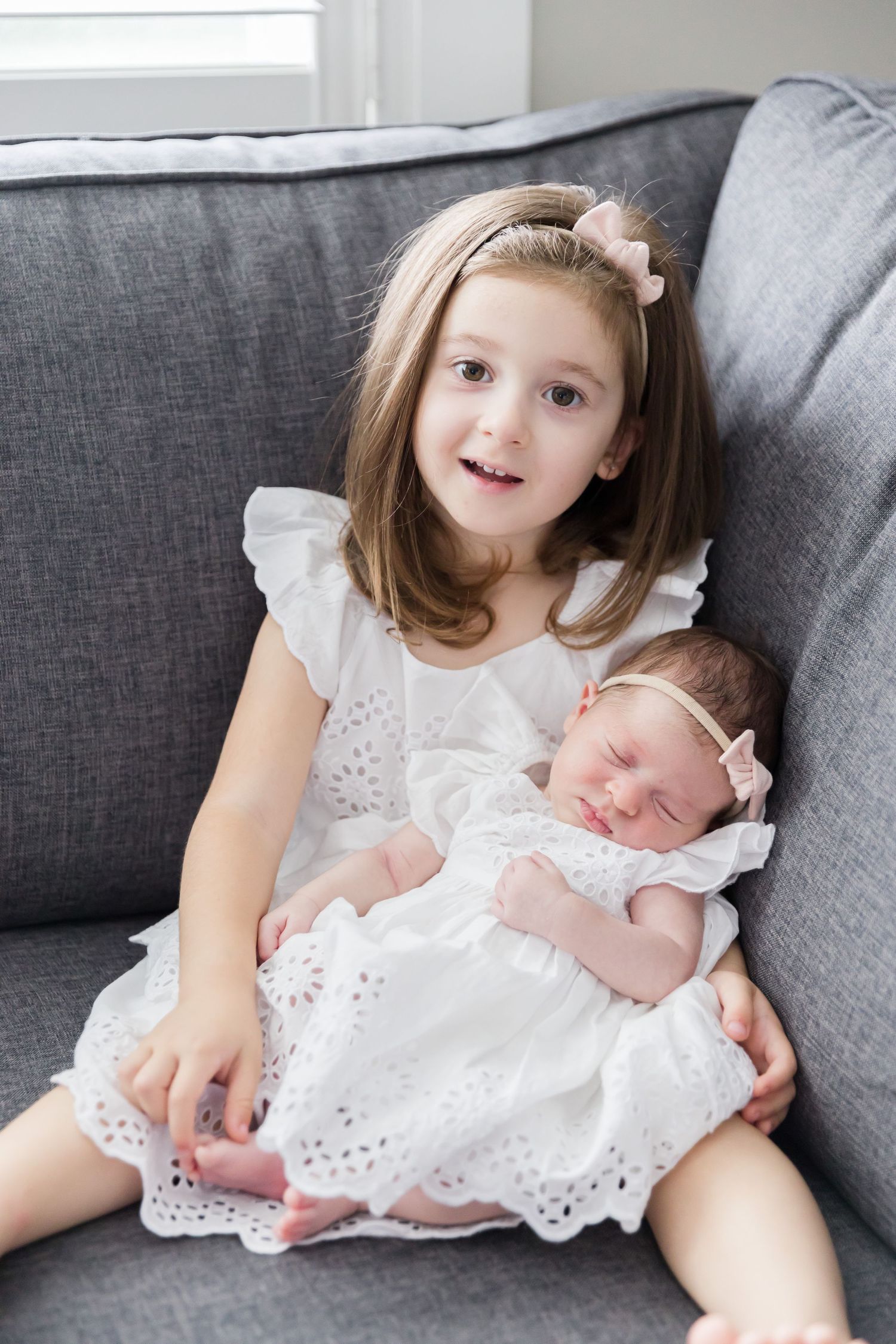 Young child holding baby sister in matching white dresses on grey sofa.