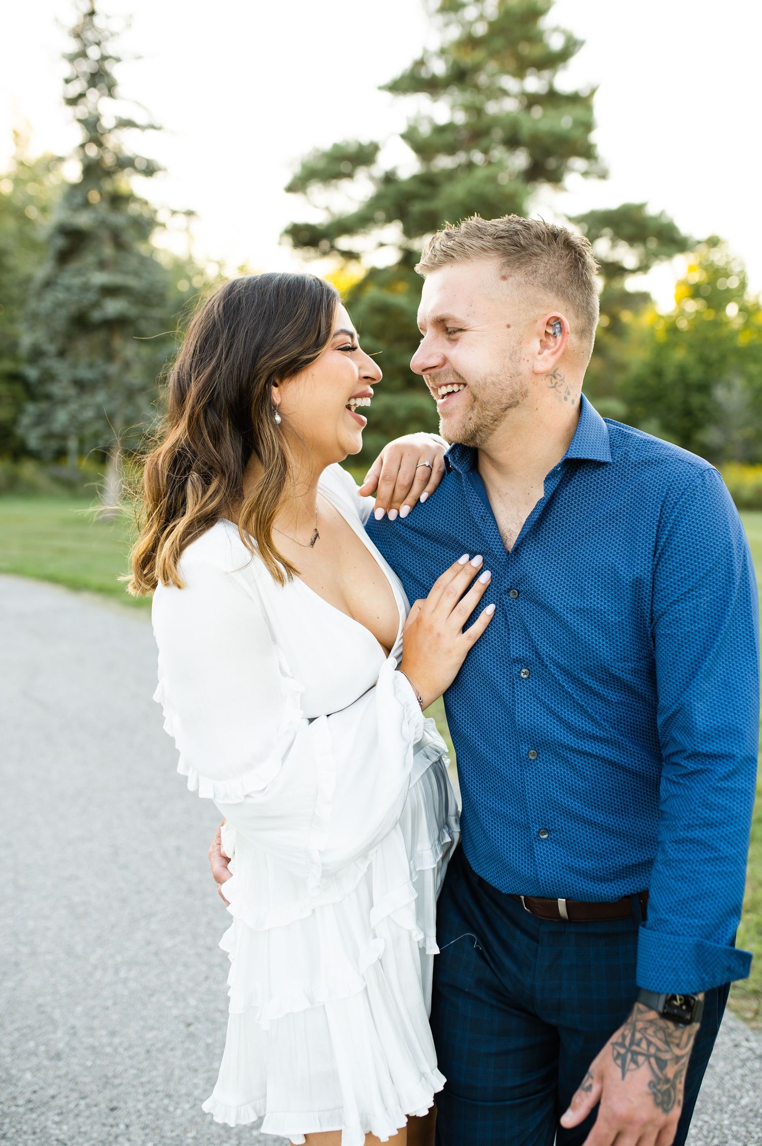 A romantic couple laughs together outdoors, wearing a white dress and blue shirt against an evergreen backdrop.